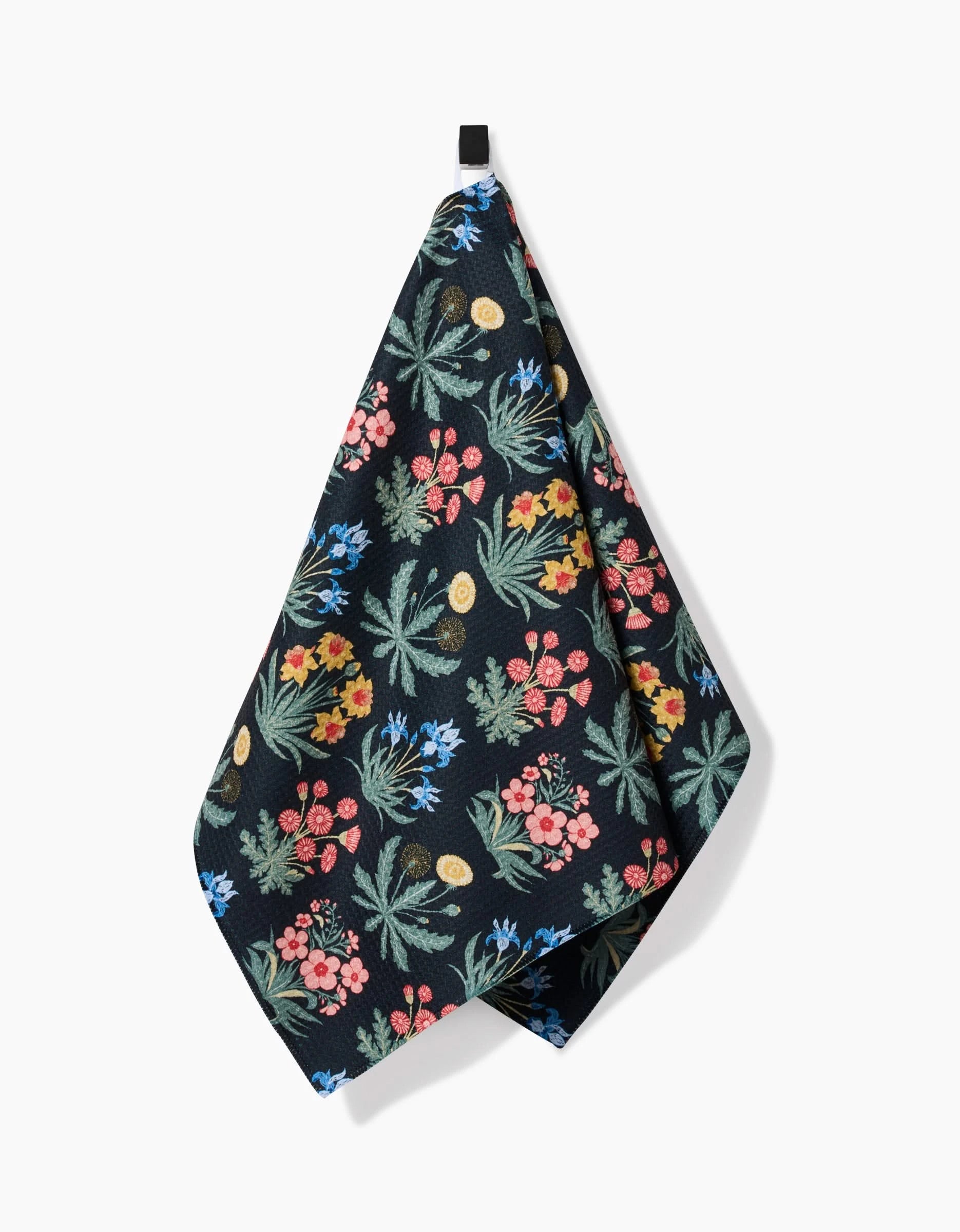 The Morning Meadow tea towel by Danelys Sidron features a vibrant pink, yellow, blue, and green floral print on black Geoweave™ fabric made from recycled materials. It hangs from a black loop against a white background.