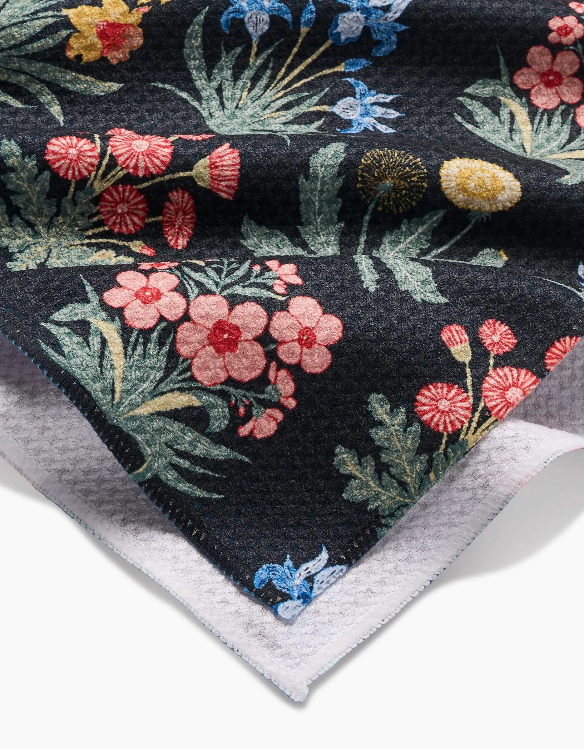 A close-up of Danelys Sidron’s Morning Meadow Tea Towel, crafted in dark Geoweave™ fabric, displays a vibrant floral pattern with red, pink, yellow, and blue flowers plus green leaves. The layered towel reveals its white underside.
