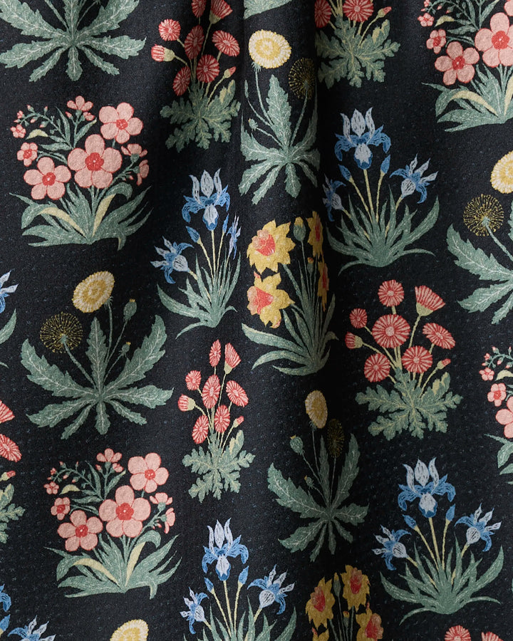 Morning Meadow by Danelys Sidron features a colorful floral pattern with clusters of pink, blue, yellow, and red flowers with green leaves on a black background. Made from recycled materials, this fabric drapes in gentle folds.