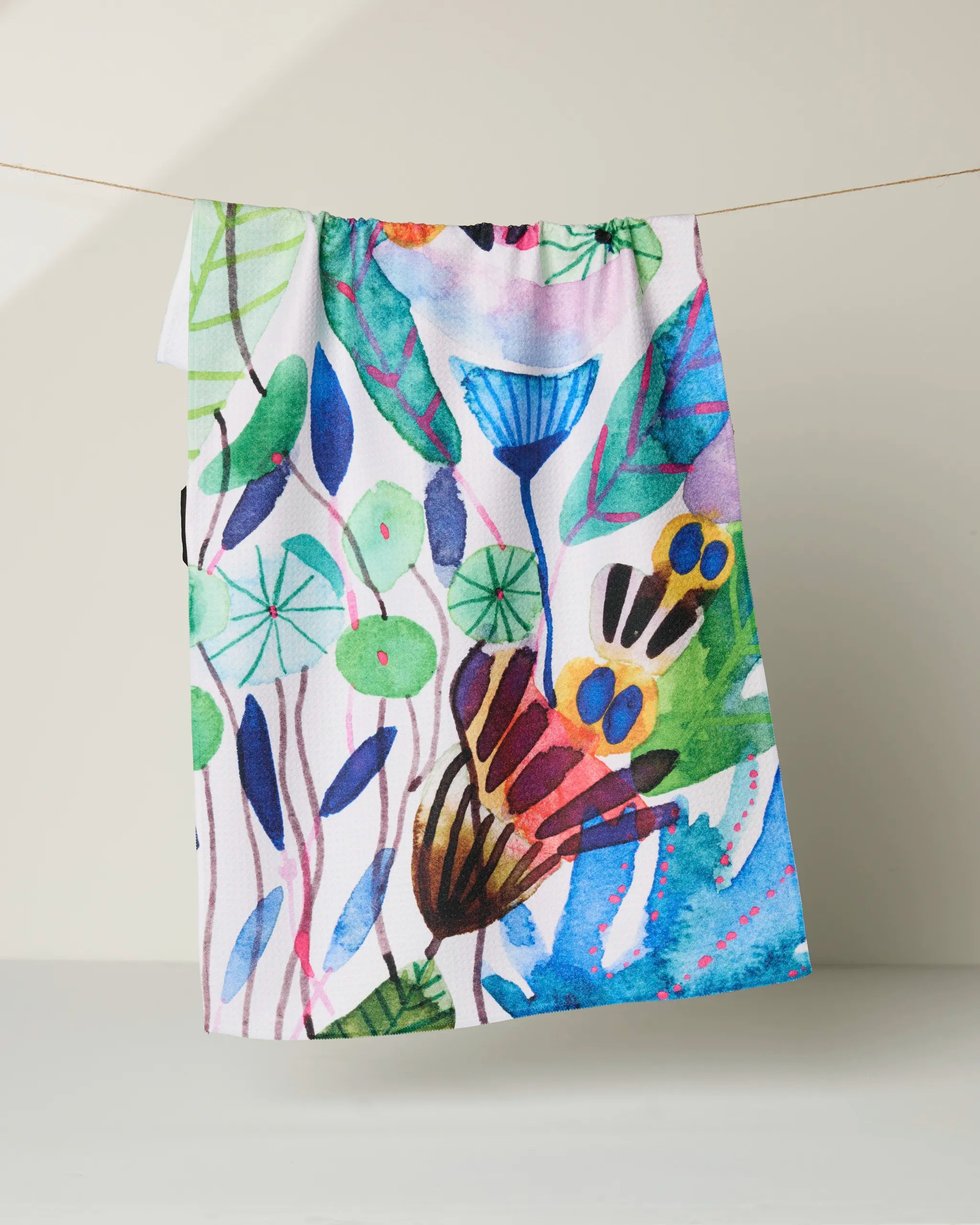 The "Morning" tea towel by Maria Montiel hangs on a string, showcasing its ultra-absorbent fabric and a vibrant watercolor pattern of leaves, flowers, and abstract shapes against a neutral backdrop.