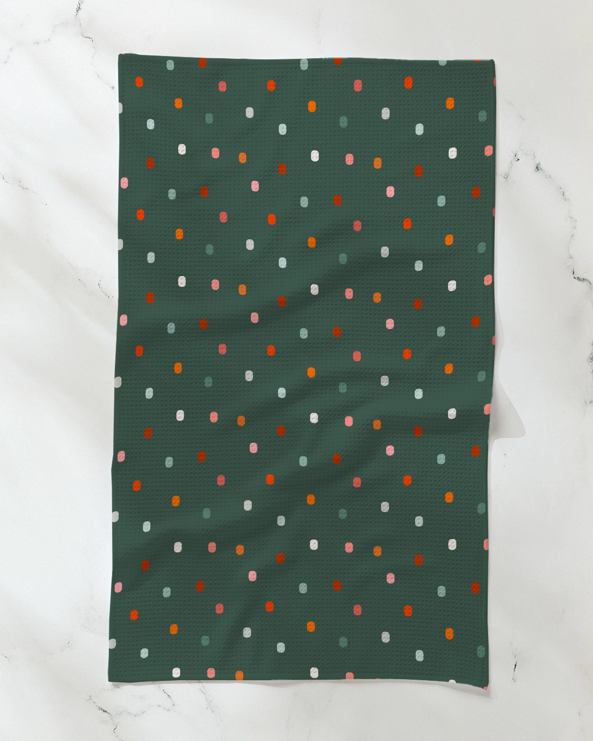 A Geometry "Moss Tile Shuffle" tea towel lies flat on a white surface, showcasing a dark green background adorned with a pattern of small, evenly spaced dots in red, pink, white, and light blue. Subtle fabric folds are visible.