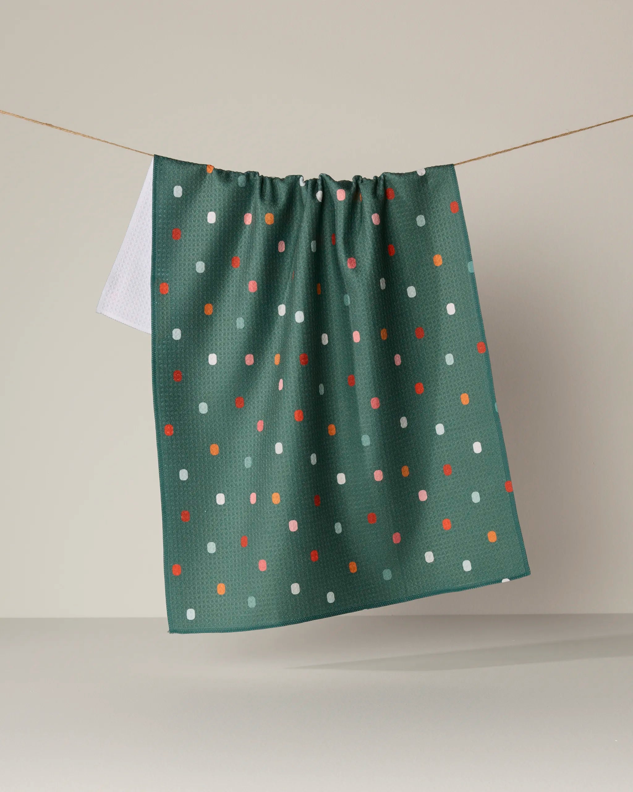 The Geometry Moss Tile Shuffle kitchen towel, featuring a vibrant polka dot pattern and crafted from recycled materials, hangs on a clothesline against a plain beige background.
