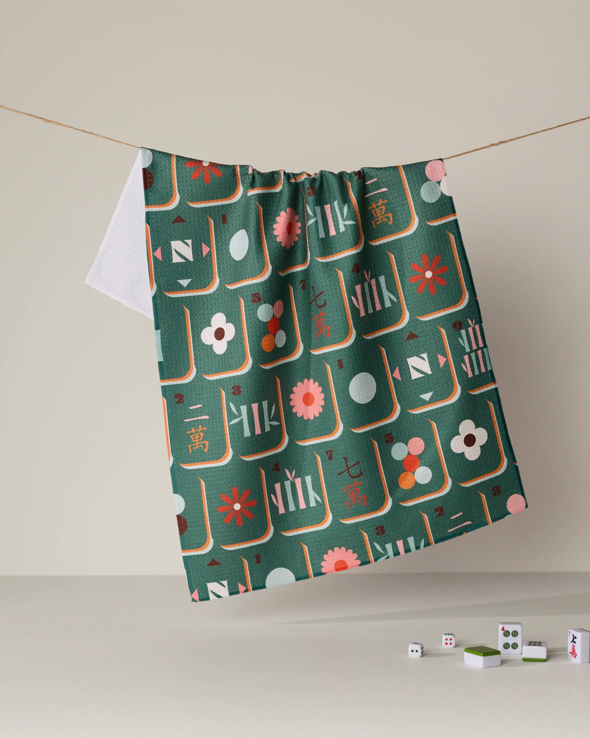 A Geometry "Moss Tile Toss" tea towel featuring Mahjong tile patterns hangs on a string, while three Mahjong tiles and two dice rest on a white surface below. This quick-drying towel combines playful charm with everyday practicality for any kitchen.