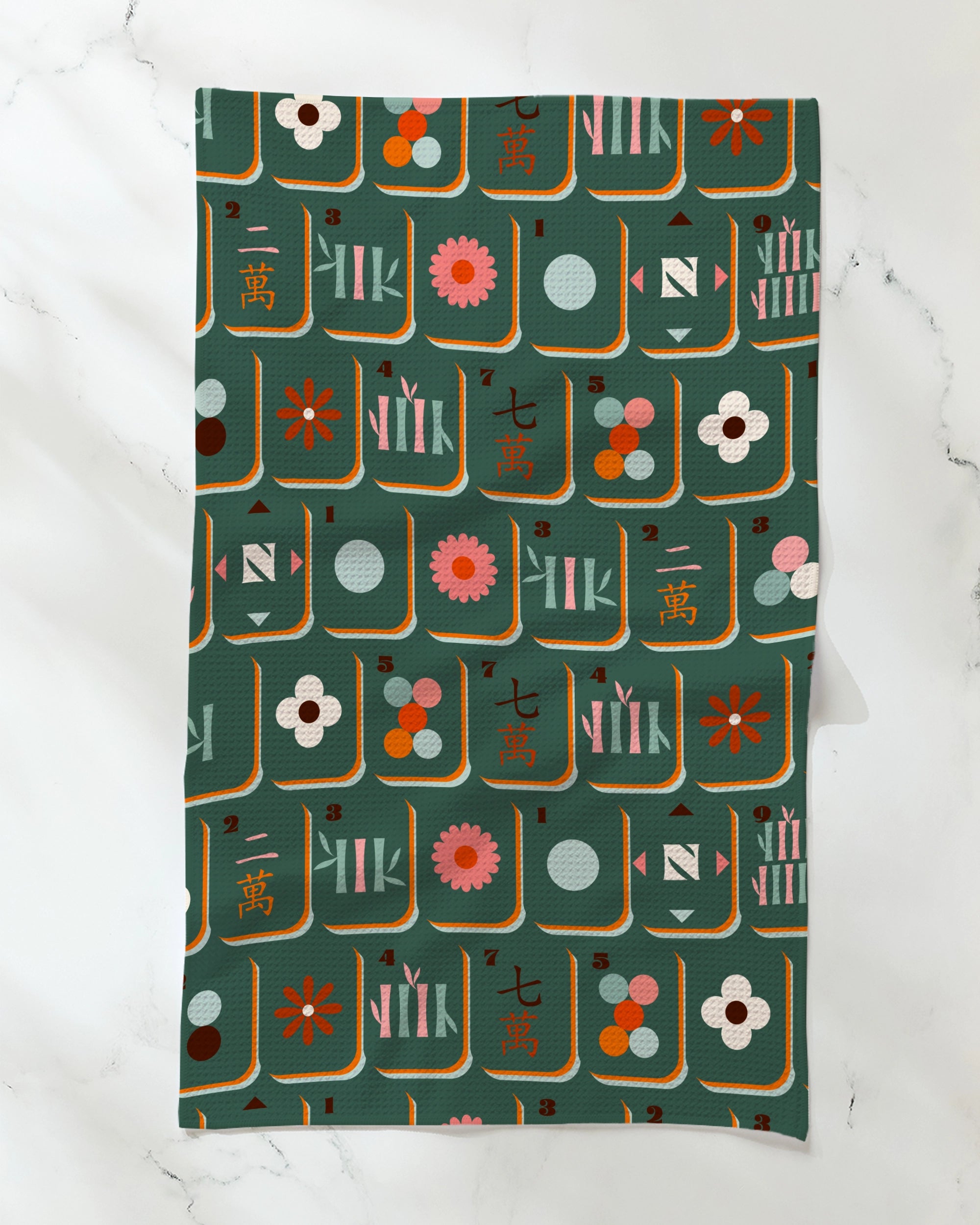 The Geometry "Moss Tile Toss" kitchen towel, crafted from recycled materials, features colorful, stylized mahjong tiles and flowers on a dark green background and is displayed on a white marble surface.