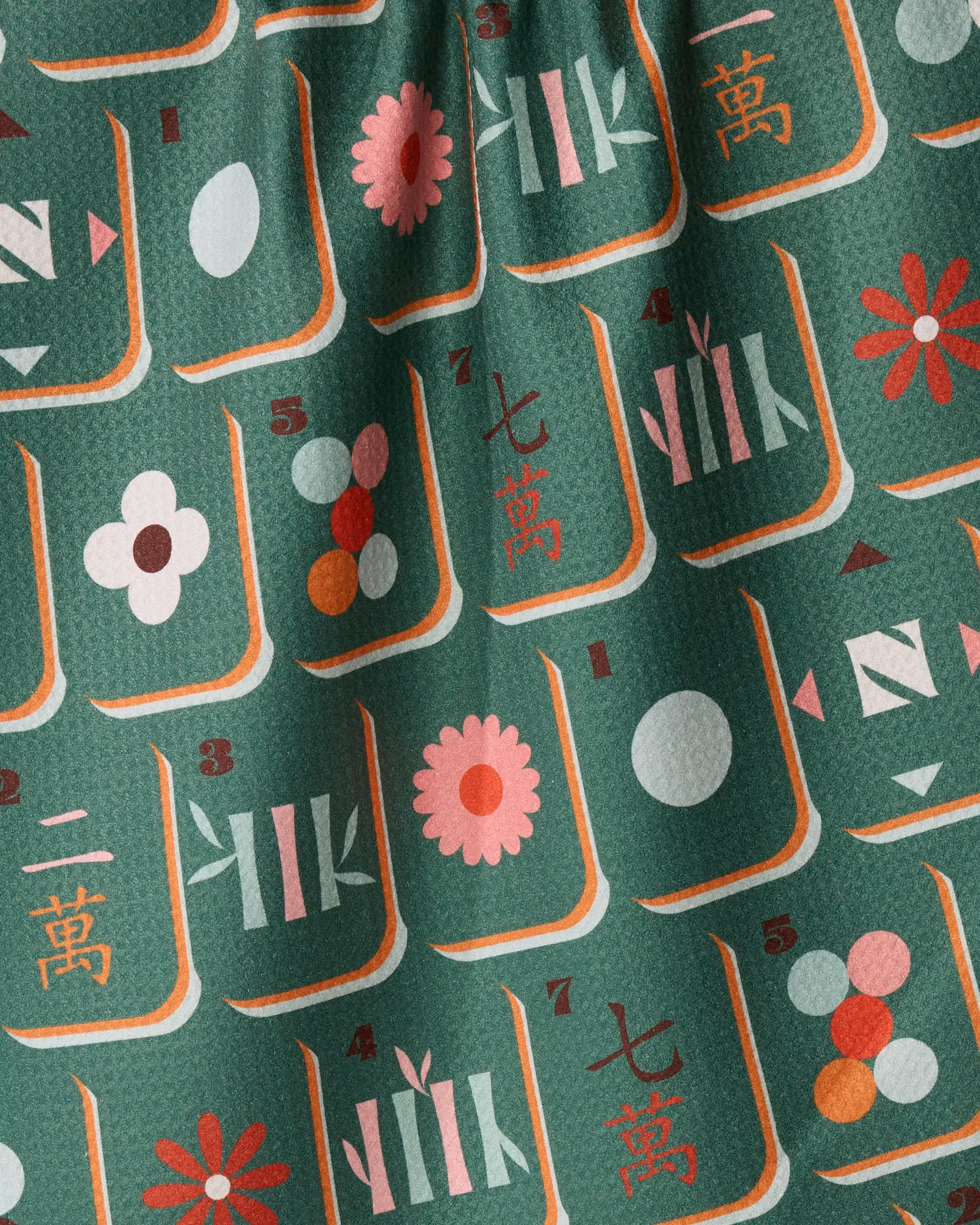 The Moss Tile Toss recycled kitchen towel by Geometry features green fabric adorned with a repeating pattern of Mahjong tiles, showcasing stylized numbers, flowers, dots, and bamboo symbols in shades of red, white, pink, and light blue. The towel has a textured surface with slight folds.