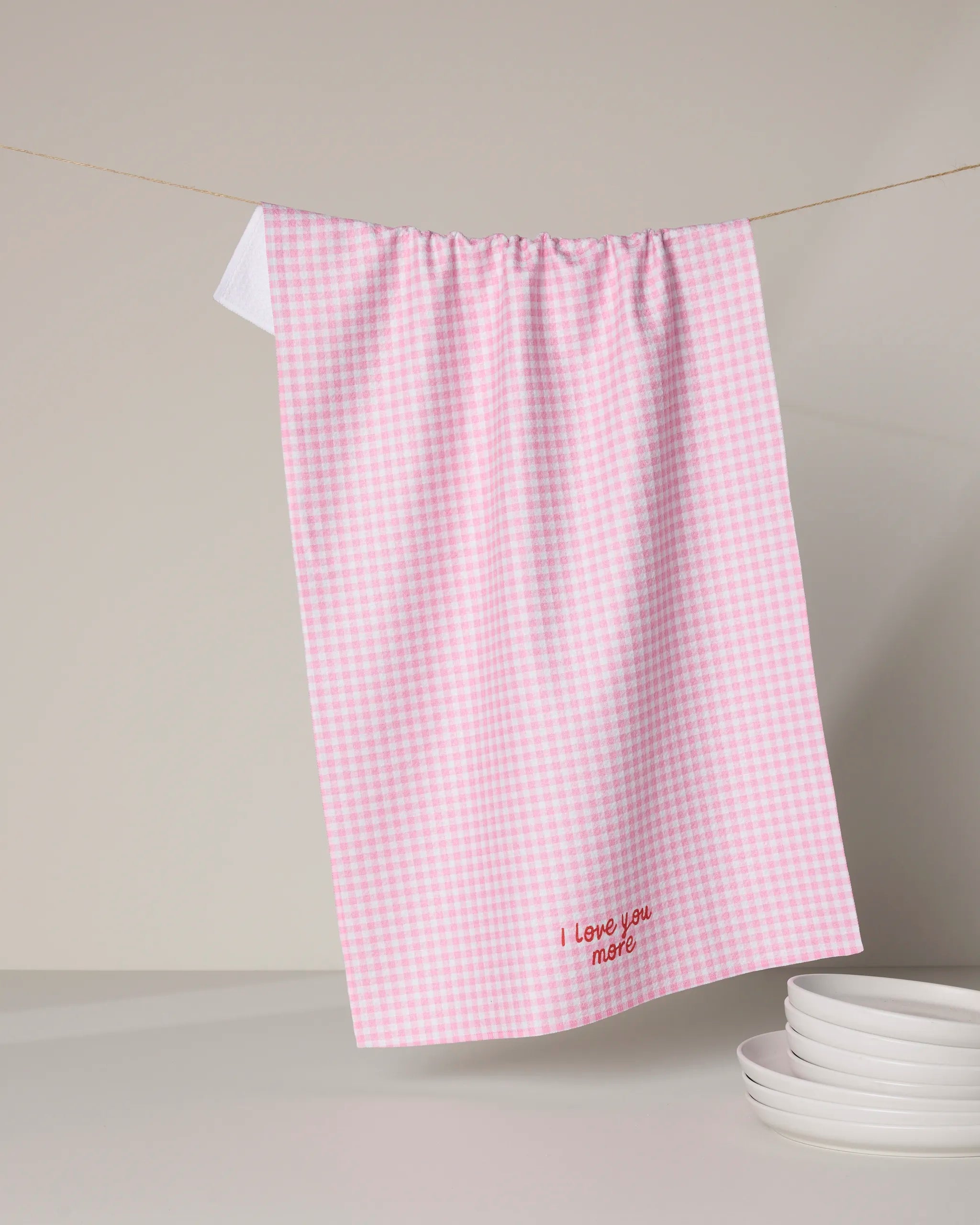 A pink and white checkered Geometry Most tea towel made from recycled materials hangs on a string, embroidered with "I love you more" at the bottom. Beneath it, several stacked white plates sit on the surface.