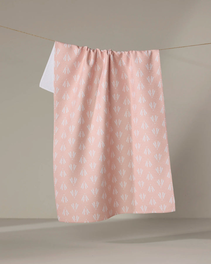 The Moth Geometric tea towel by Hufton Studio, featuring a white arrow pattern on light pink Geoweave fabric, hangs on a clothesline against a neutral beige background.