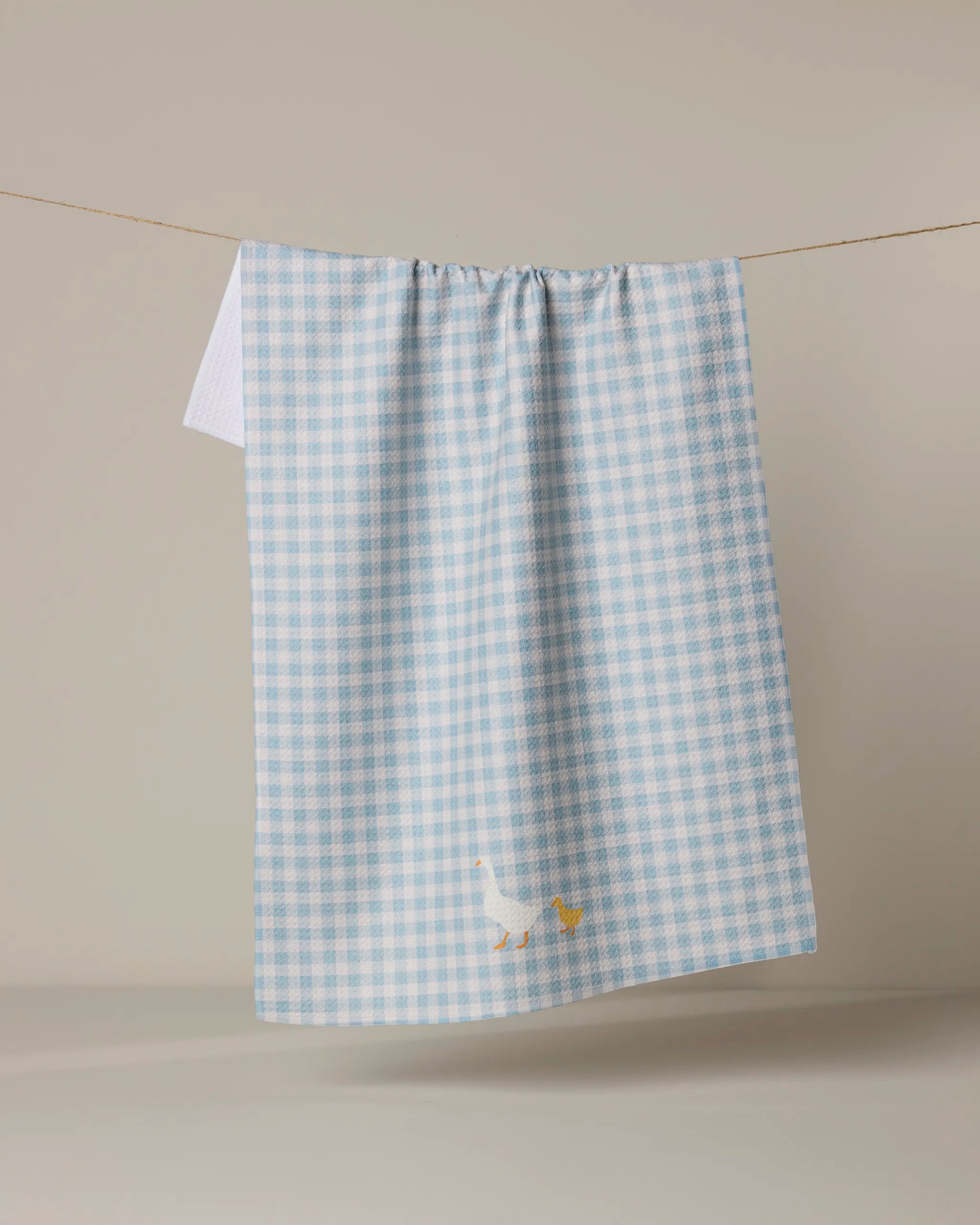 The Geometry Mother Alli is a blue and white checkered absorbent dish towel featuring an embroidered white duck near the bottom, shown hanging on a clothesline against a plain beige background.