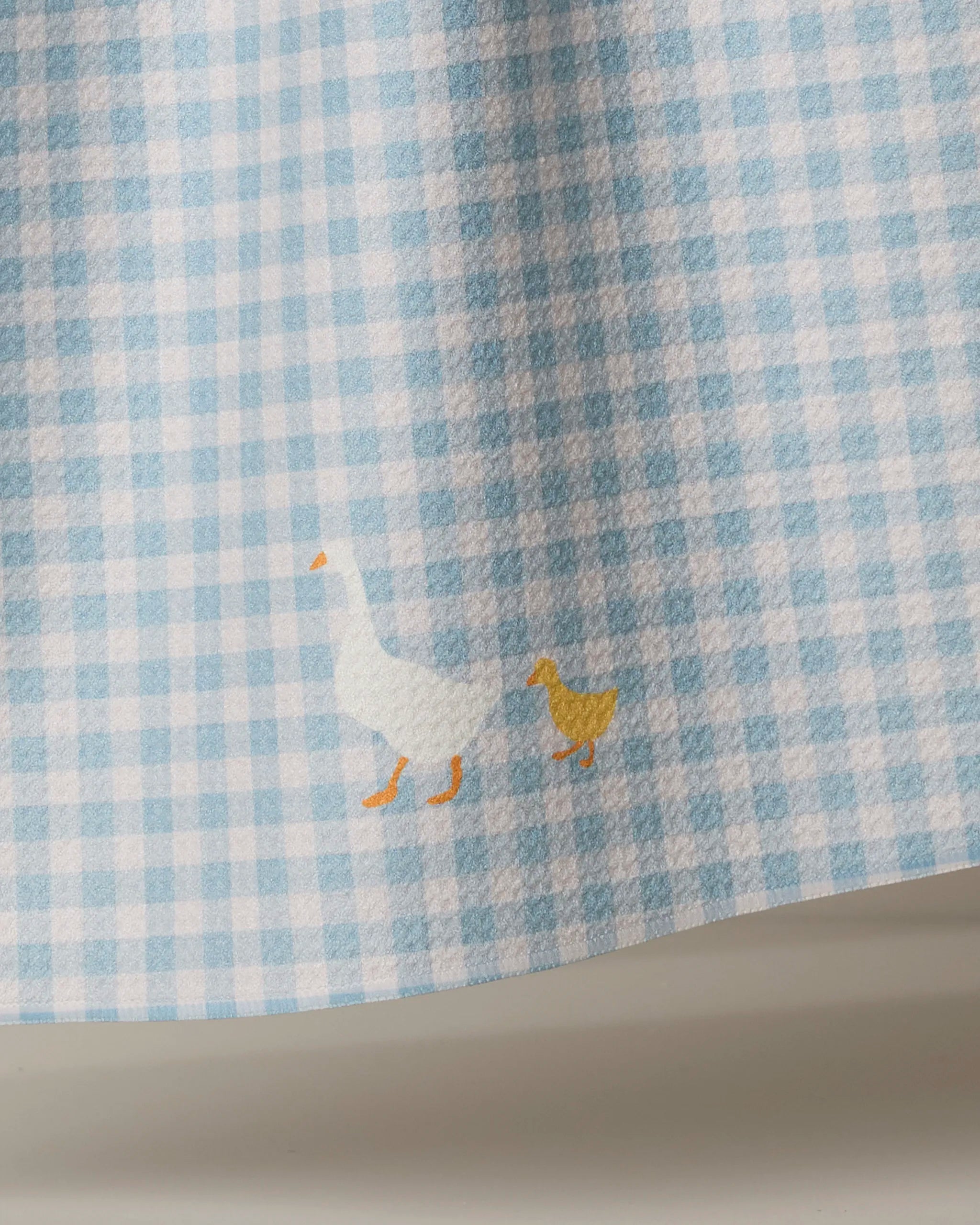 The Geometry Mother Alli dish towel is blue and white gingham, featuring a small illustration near the bottom edge of a white goose and yellow gosling walking side by side.