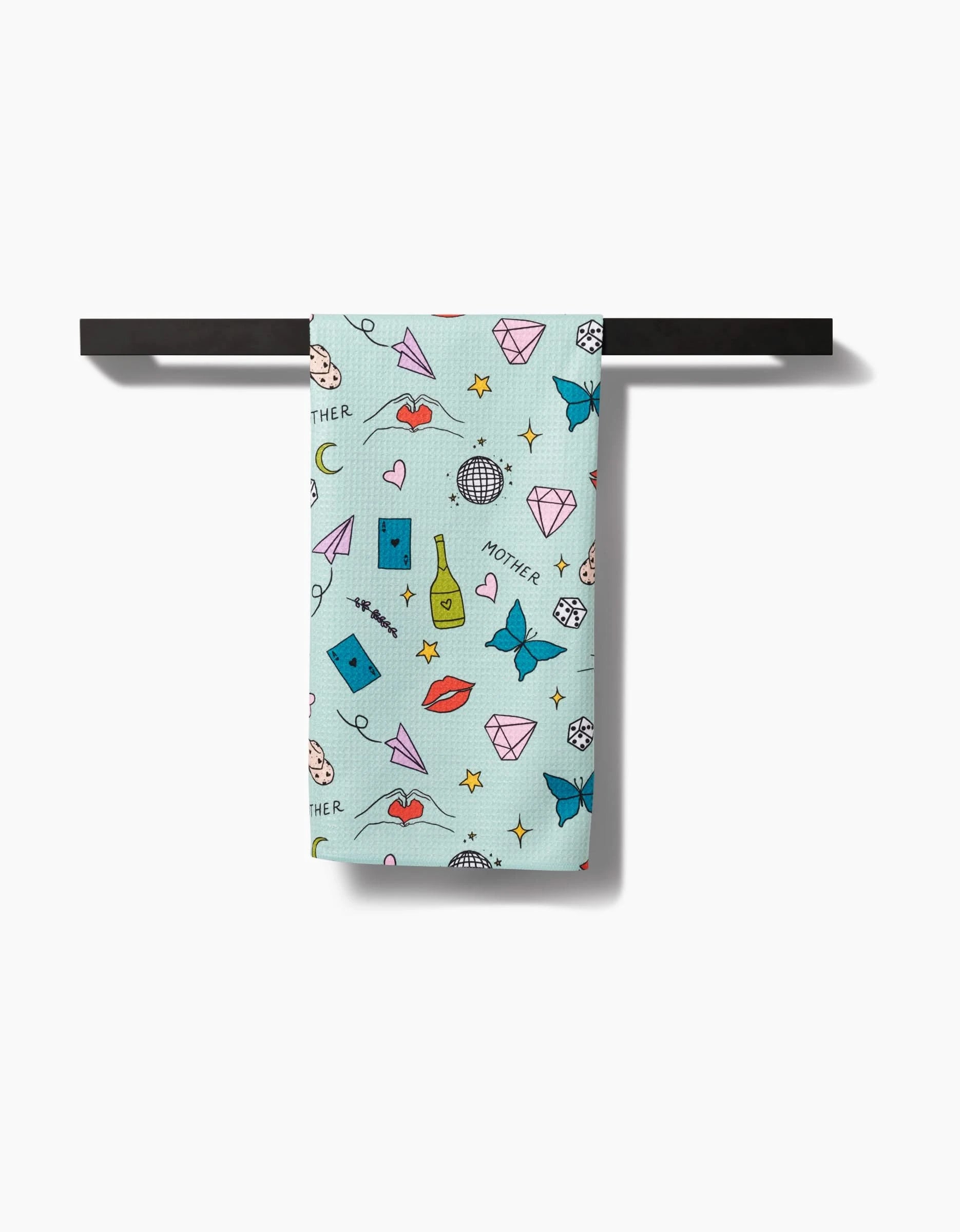 The Mother towel by Sarah Holden is a light blue towel made from recycled materials, featuring colorful illustrations and the word MOTHER. Its shown hanging on a black towel bar.