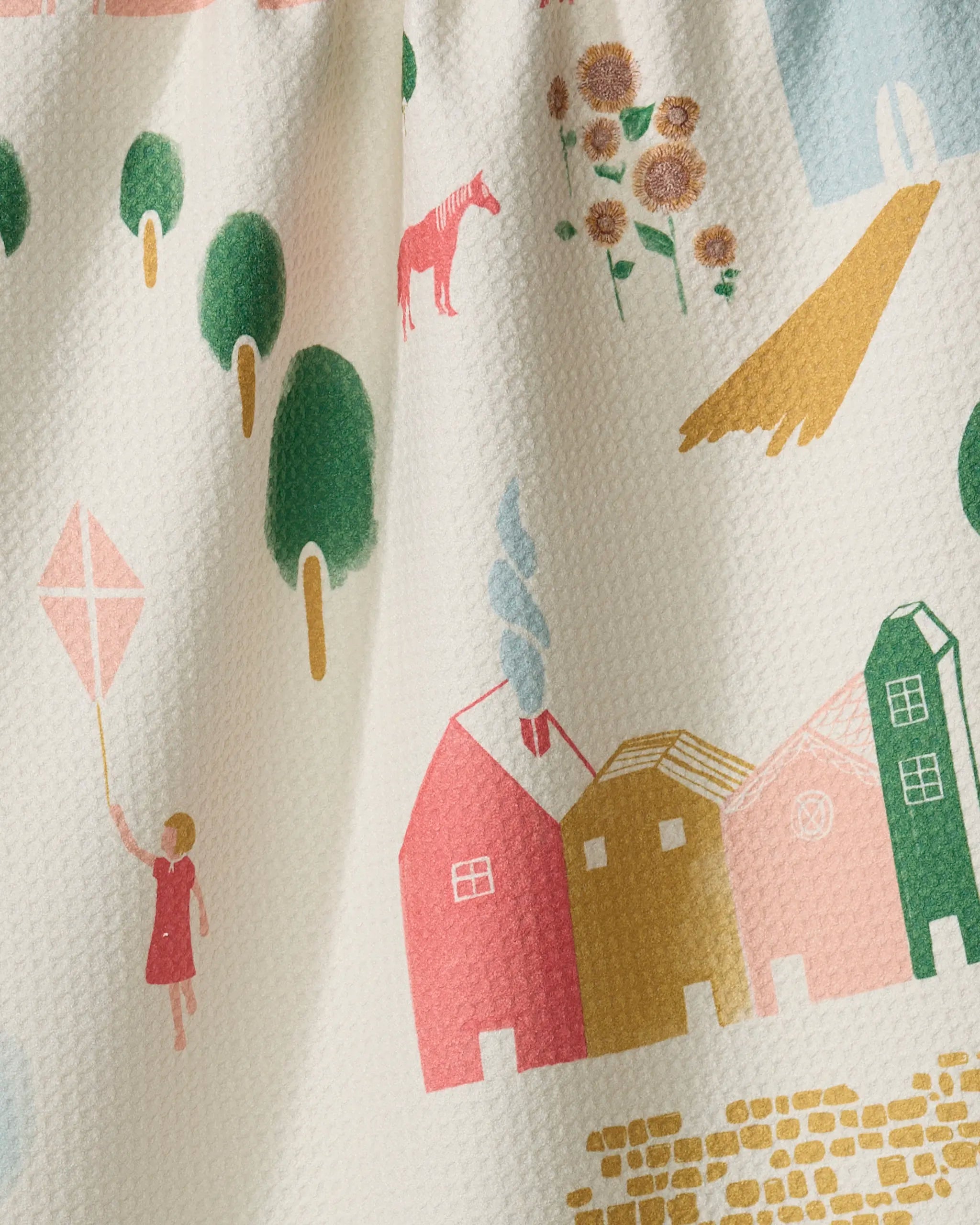 Motherland by Elisabeth Wing: a whimsical illustration on textured fabric featuring colorful houses, trees, sunflowers, a horse, and a person flying a pink kite—ideal for an eco-friendly kitchen towel.