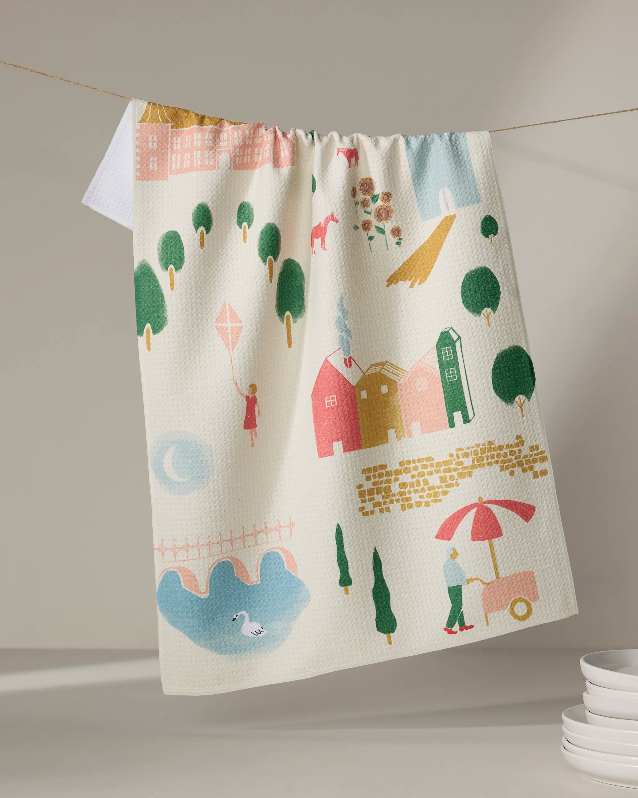 Motherland by Elisabeth Wing is a colorful tea towel made from recycled materials, depicting whimsical houses, trees, a bridge with a swan, people strolling, kite flyers, and an ice cream cart under the sun. Plates are stacked nearby.