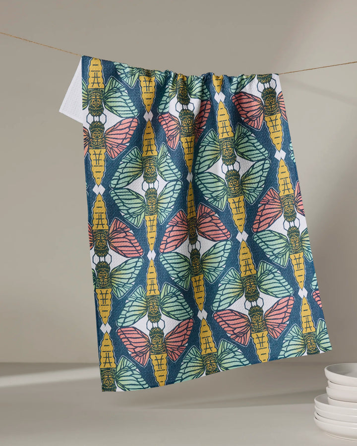The Renee Fly "Moths" tea towel, featuring a vibrant pink, yellow, and green moth pattern, dries quickly on the line beside stacked white plates on a light surface.