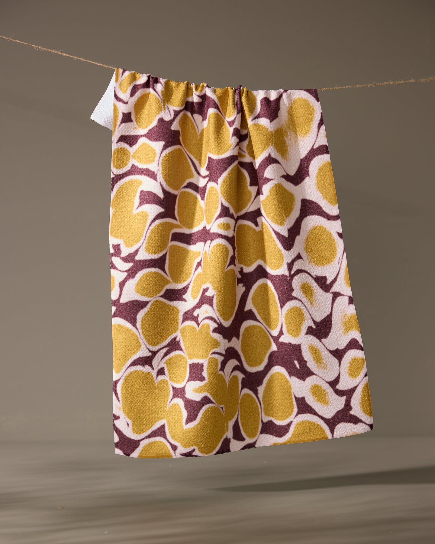 The Geometry Mottled recycled kitchen towel, featuring a yellow and brown abstract pattern, hangs from a string against a neutral background.