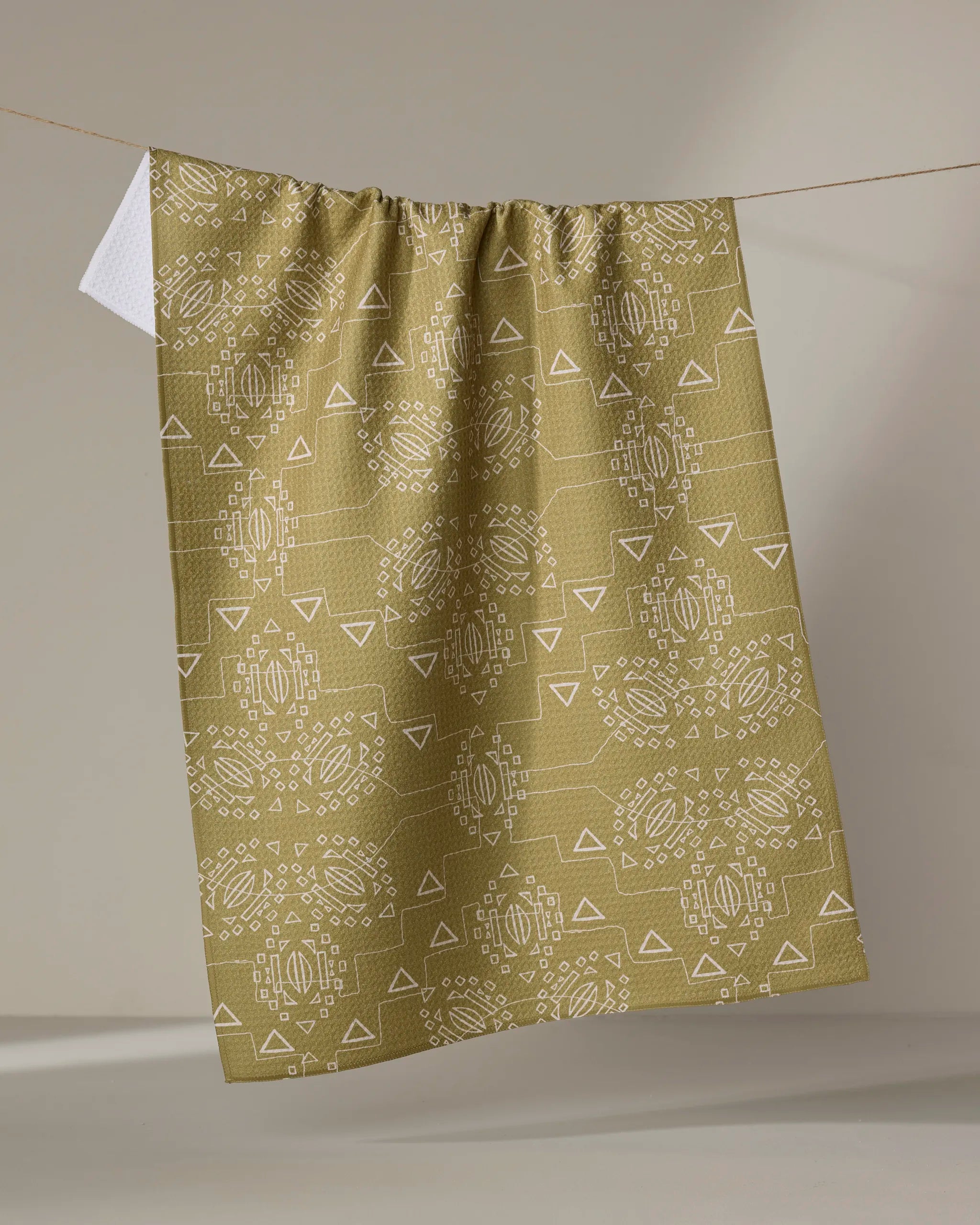 The Ash Cascade "Mountain Bloom" tea towel, crafted from recycled materials in light brown with white geometric and circular designs, hangs on a line showing its crisp white reverse against a neutral background.
