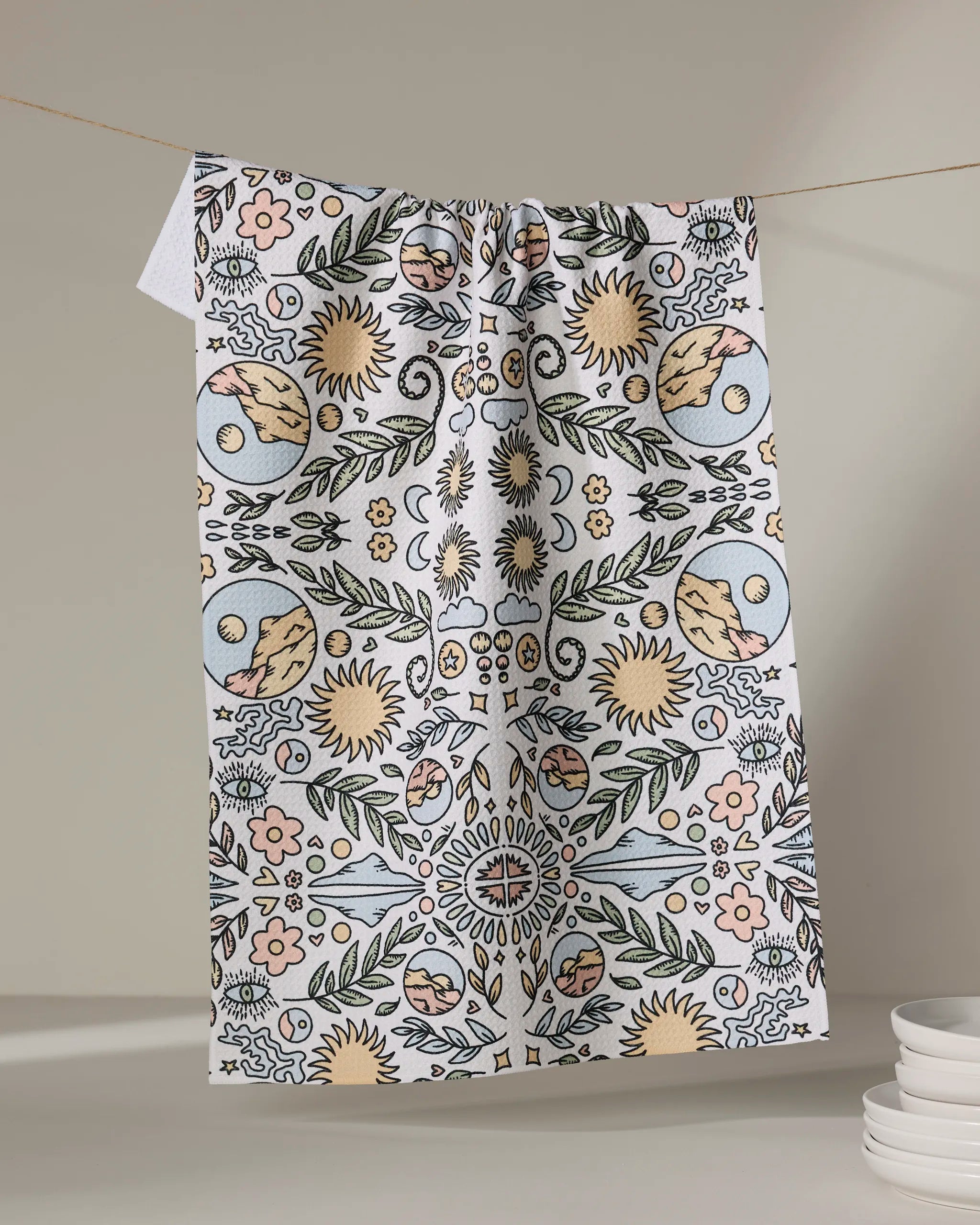 The Mountain Mosaic tea towel by Graphics and Grain, featuring sun, moon, and floral pastel designs, hangs on a clothesline near a stack of white plates against a neutral wall.