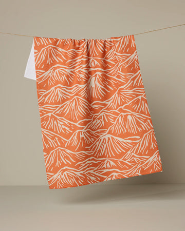 The Mountain Range tea towel by Geometry, featuring an orange and white mountain pattern, hangs from a string against a plain beige background.