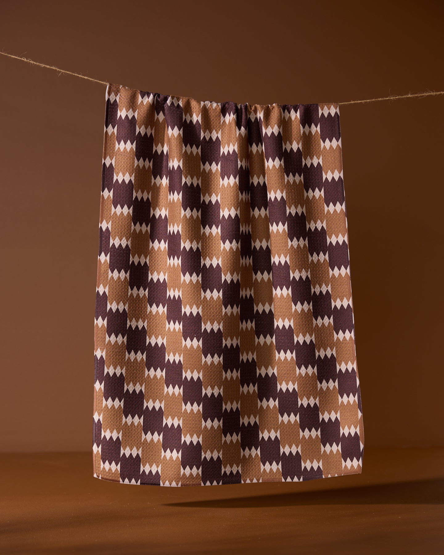 The Geometry Mountain Song recycled kitchen towel, featuring a geometric pattern in brown, cream, and dark purple, hangs against a plain brown background.