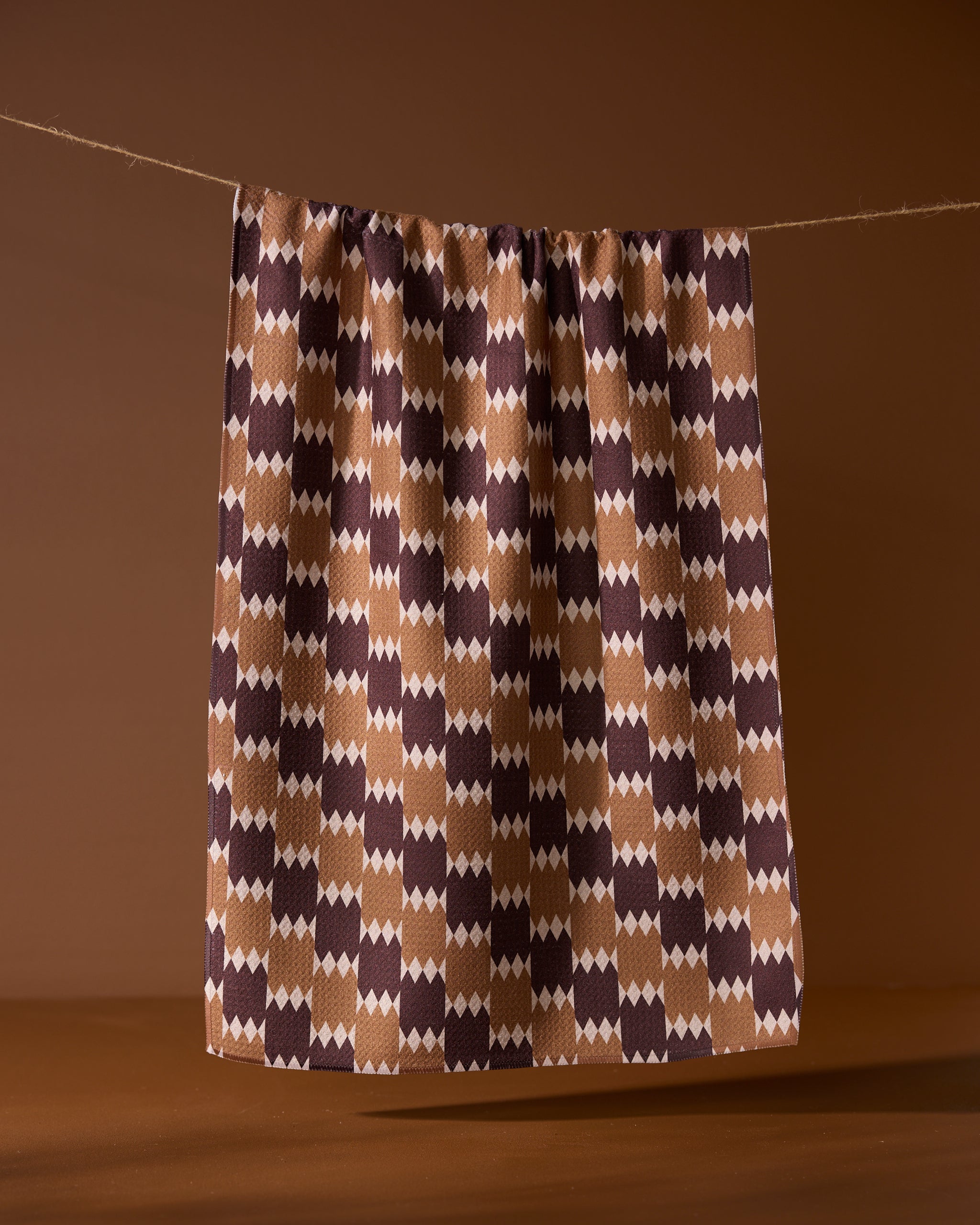 The Geometry Mountain Song recycled kitchen towel, featuring a geometric pattern in brown, cream, and dark purple, hangs against a plain brown background.