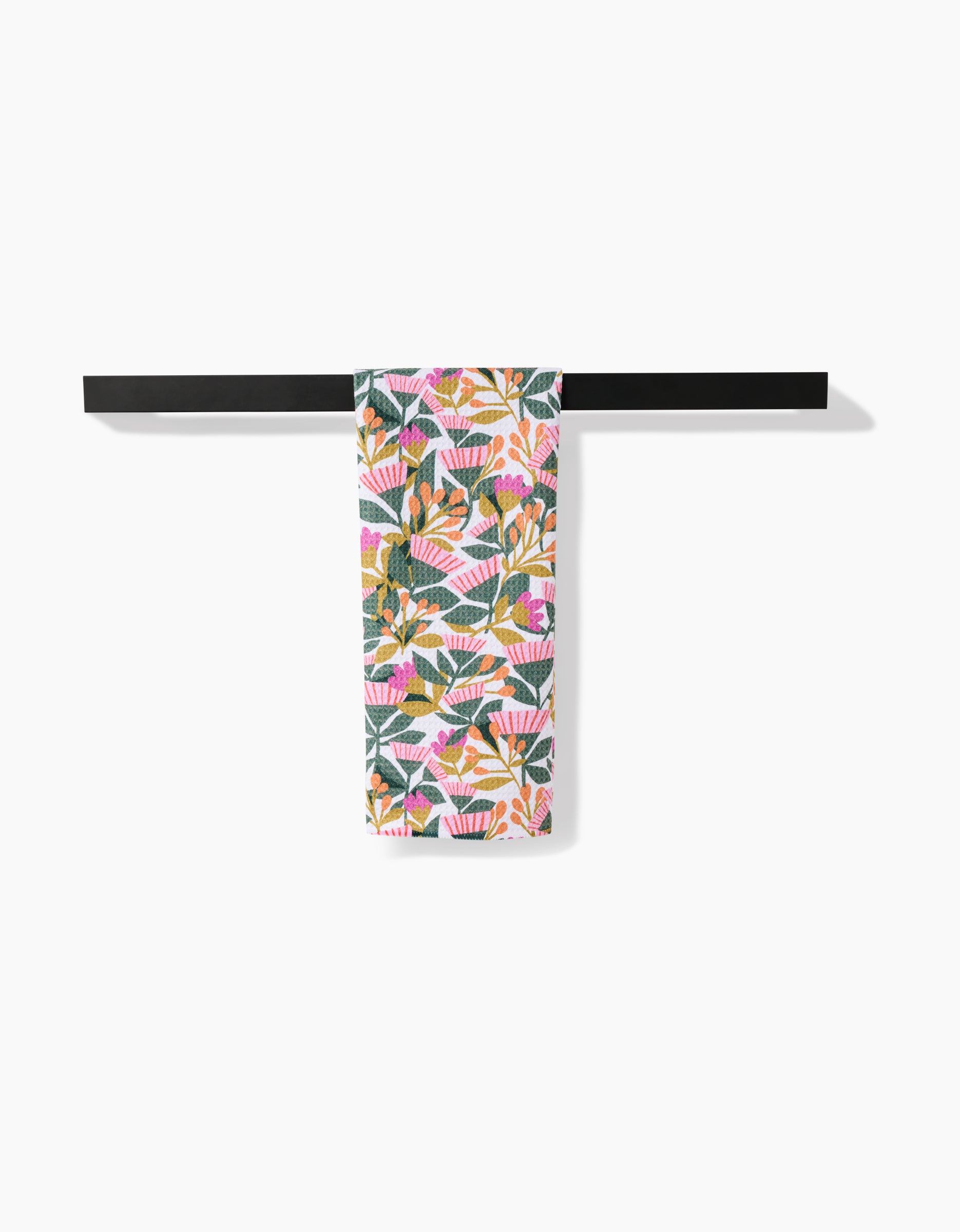 The Moxie Mauve tea towel by Lisa Nelson, featuring a vibrant tropical leaf and flower pattern in pink, orange, green, and yellow recycled fabric, hangs neatly over a sleek black towel bar against a white background.
