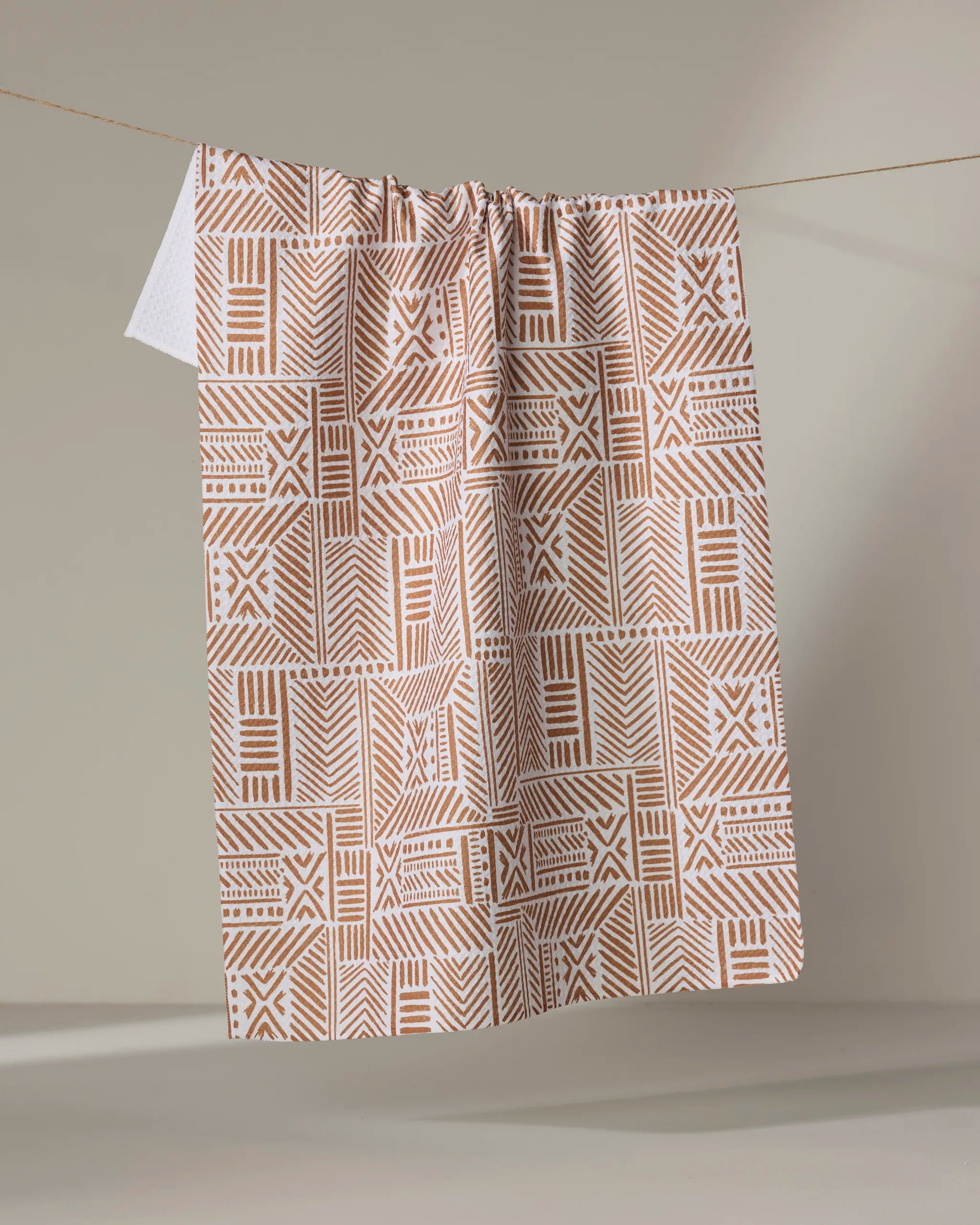 The Mud Cloth Natural kitchen towel by Hufton Studio, featuring geometric and tribal-inspired beige and white designs, is crafted from recycled materials and hangs over a clothesline against a neutral background.
