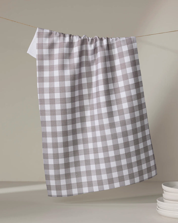 A chocolate and white checkered recycled kitchen towel by The Buy Guide (Multi Gingham - Chocolate) hangs on a clothesline, with a stack of white plates partially visible in the lower right corner against a neutral background.