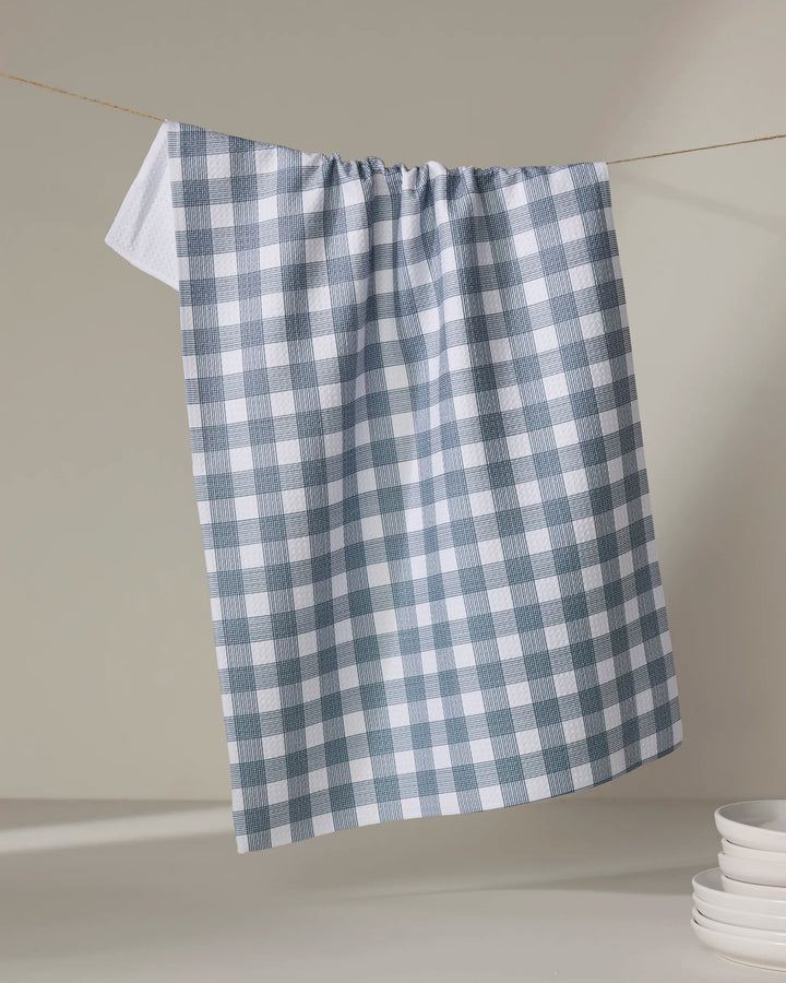 A Multi Gingham - Navy kitchen towel by The Buy Guide hangs on a clothesline, with a stack of white plates partially visible in the lower right corner against a neutral background.
