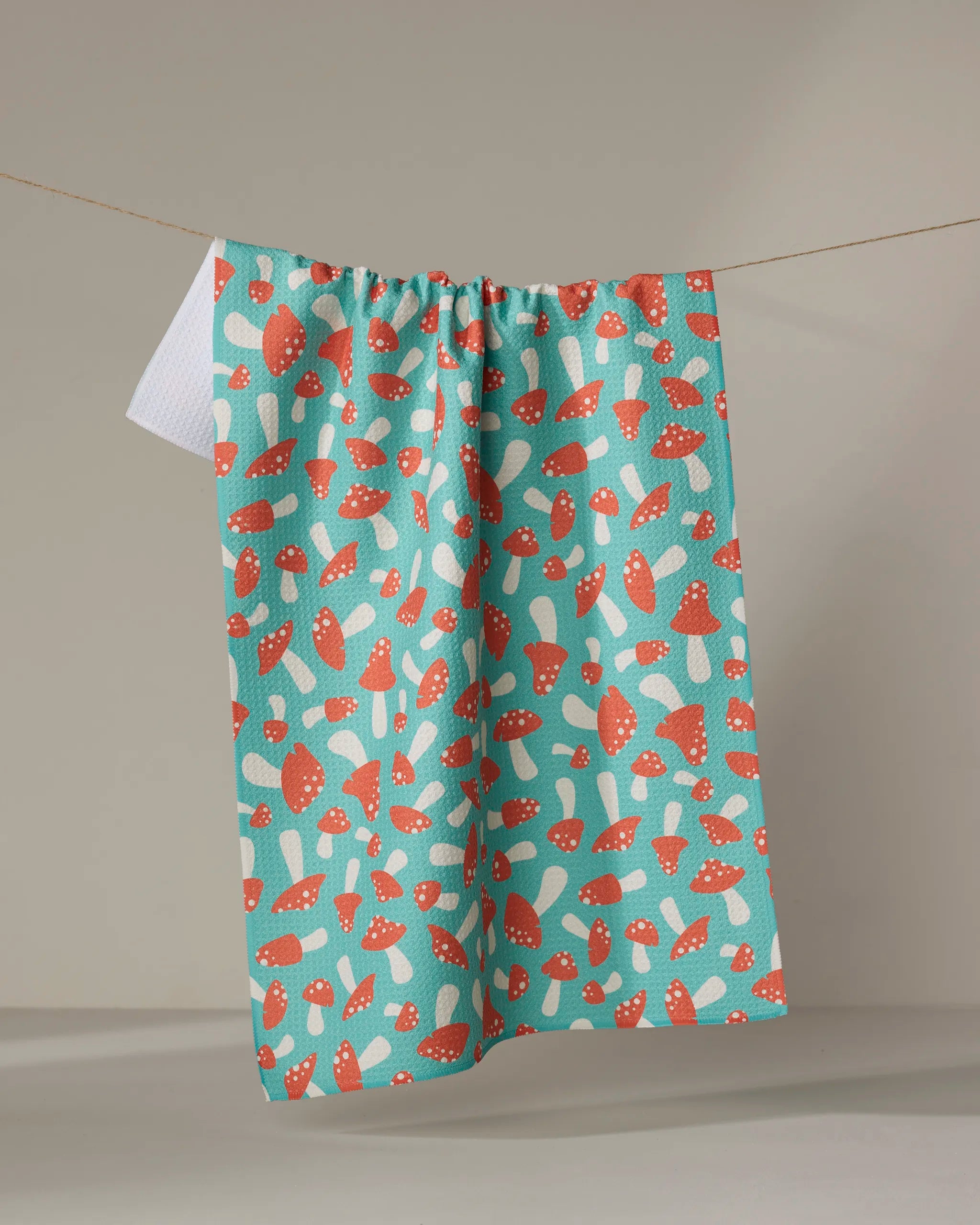 The Mushrooms towel by Megan Kampa features a playful red and white mushroom pattern and quick-drying fabric, shown hanging on a clothesline against a neutral backdrop.