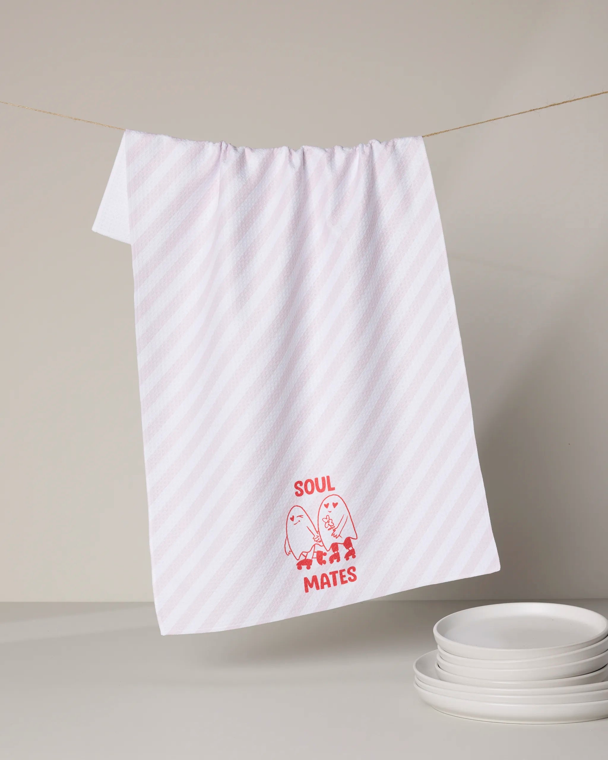 A white tea towel from Geometry, called My Ghostly Love, hangs on a line. Made from recycled materials, it features a red graphic of two smiling eggs holding hands with "SOUL MATES" below. White plates are stacked nearby.