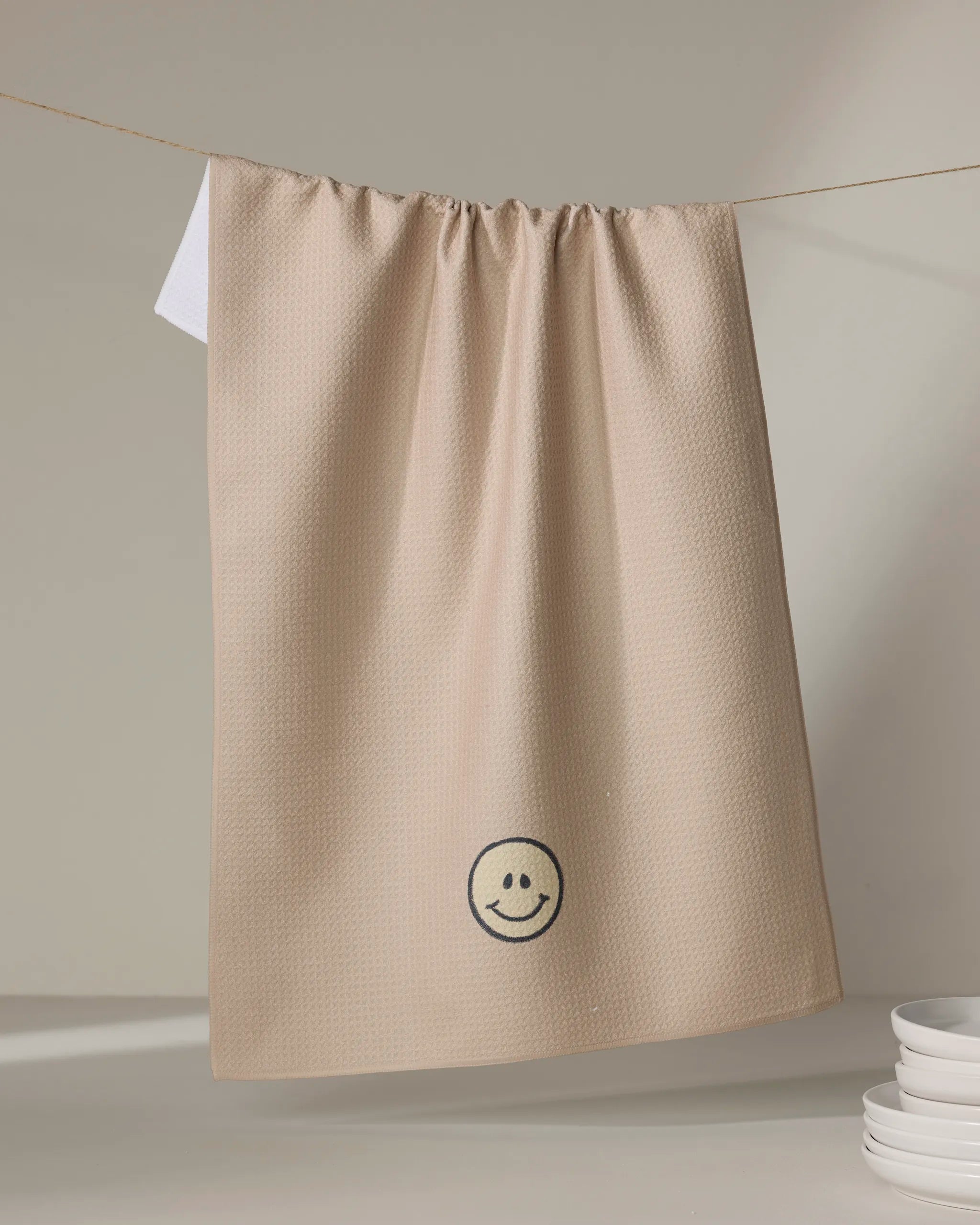 The "My Happy Place" kitchen towel by Kelsi Fullmer, featuring a beige textured design and an embroidered black smiley face, hangs on a clothesline above stacked white dishes, adding charm to your eco-friendly kitchen.