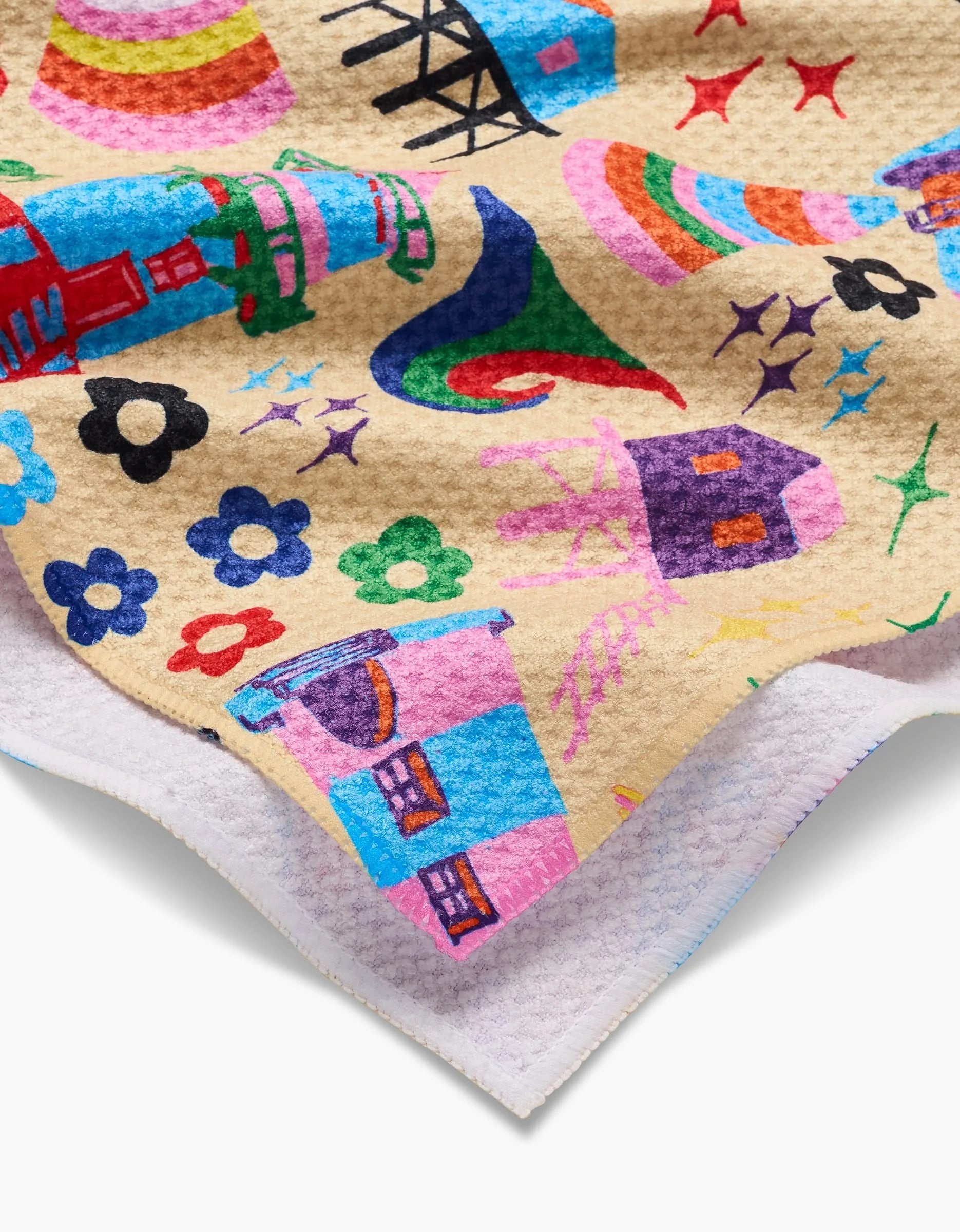 A close-up of My Place by Ania Zwara Art—a colorful tea towel made from recycled materials, featuring whimsical cartoon houses, stars, flowers, and swirls on a tan background with a white border.