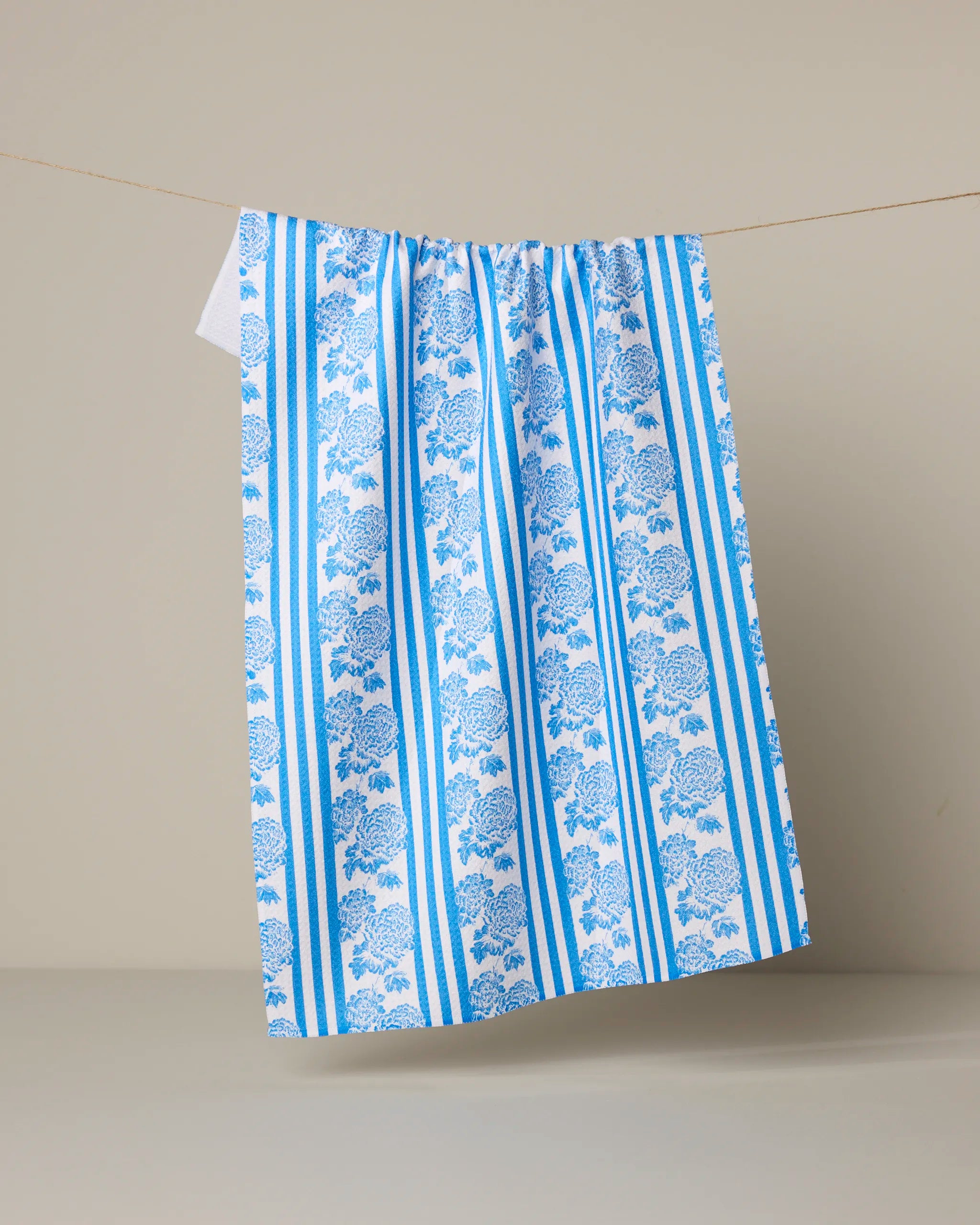 The Geometry Mykonos tea towel, featuring blue and white stripes with a floral pattern and made from recycled materials, hangs from a thin line against a plain beige background.