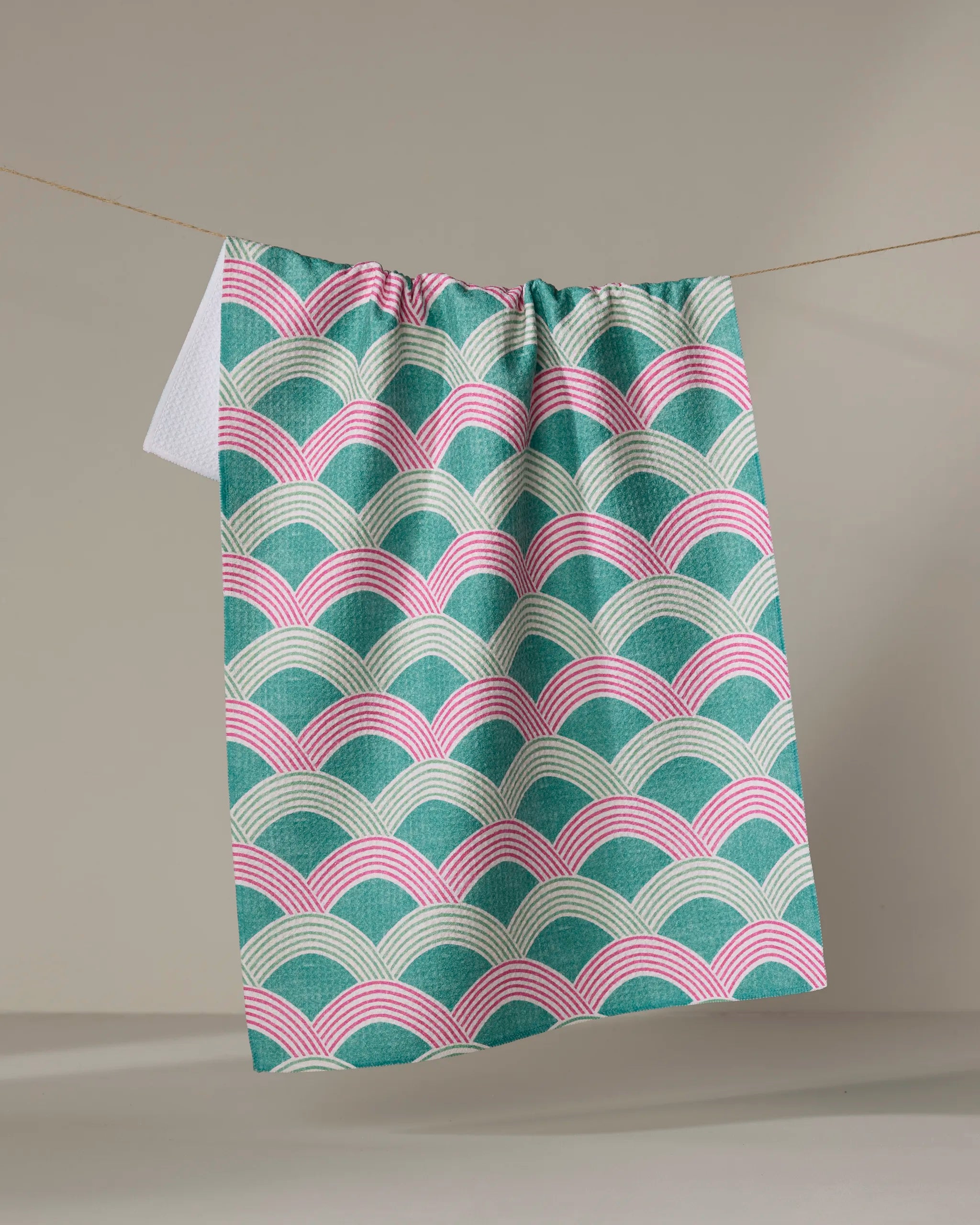 The Nalin tea towel by Amy MacCready features a scalloped teal, pink, and white pattern. Made from ultra-absorbent recycled materials, it’s shown hanging on a clothesline against a plain beige background.