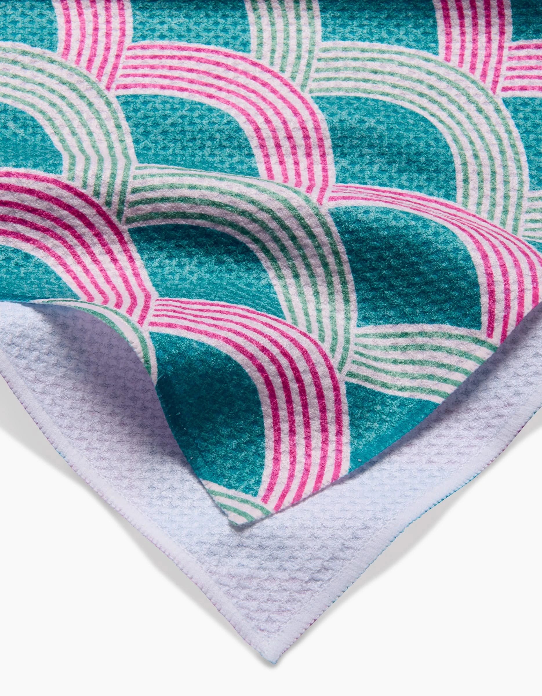 A close-up of the Amy MacCready Nalin tea towel showcases its Geoweave fabric and geometric pattern with overlapping teal, pink, and white curves. A folded corner reveals the light blue underside, crafted from recycled materials.