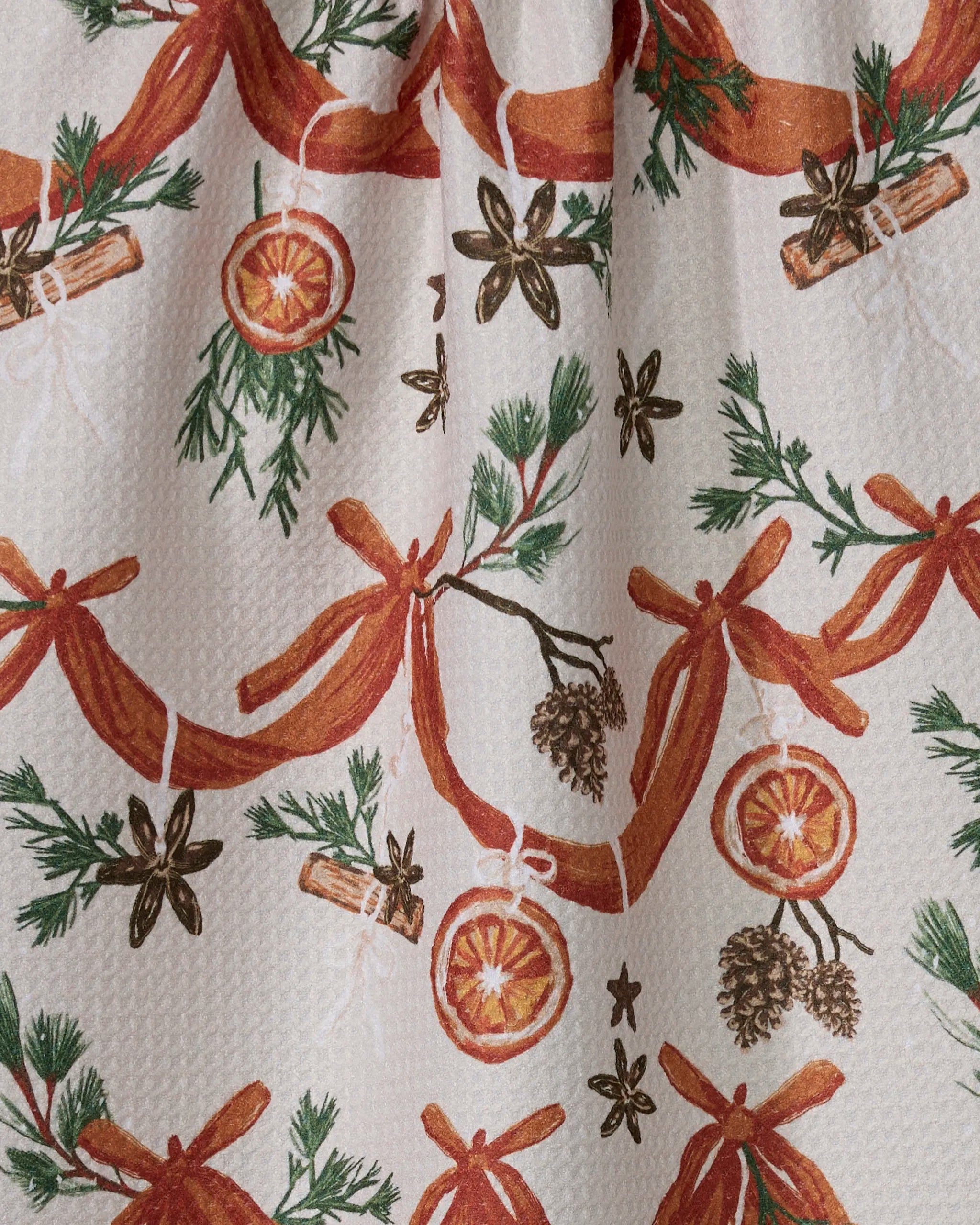 The Geometry Naranjas recycled kitchen towel features a festive cream background with a repeating pattern of orange ribbons, pine branches with cones, dried orange slices, cinnamon sticks, and star anise.