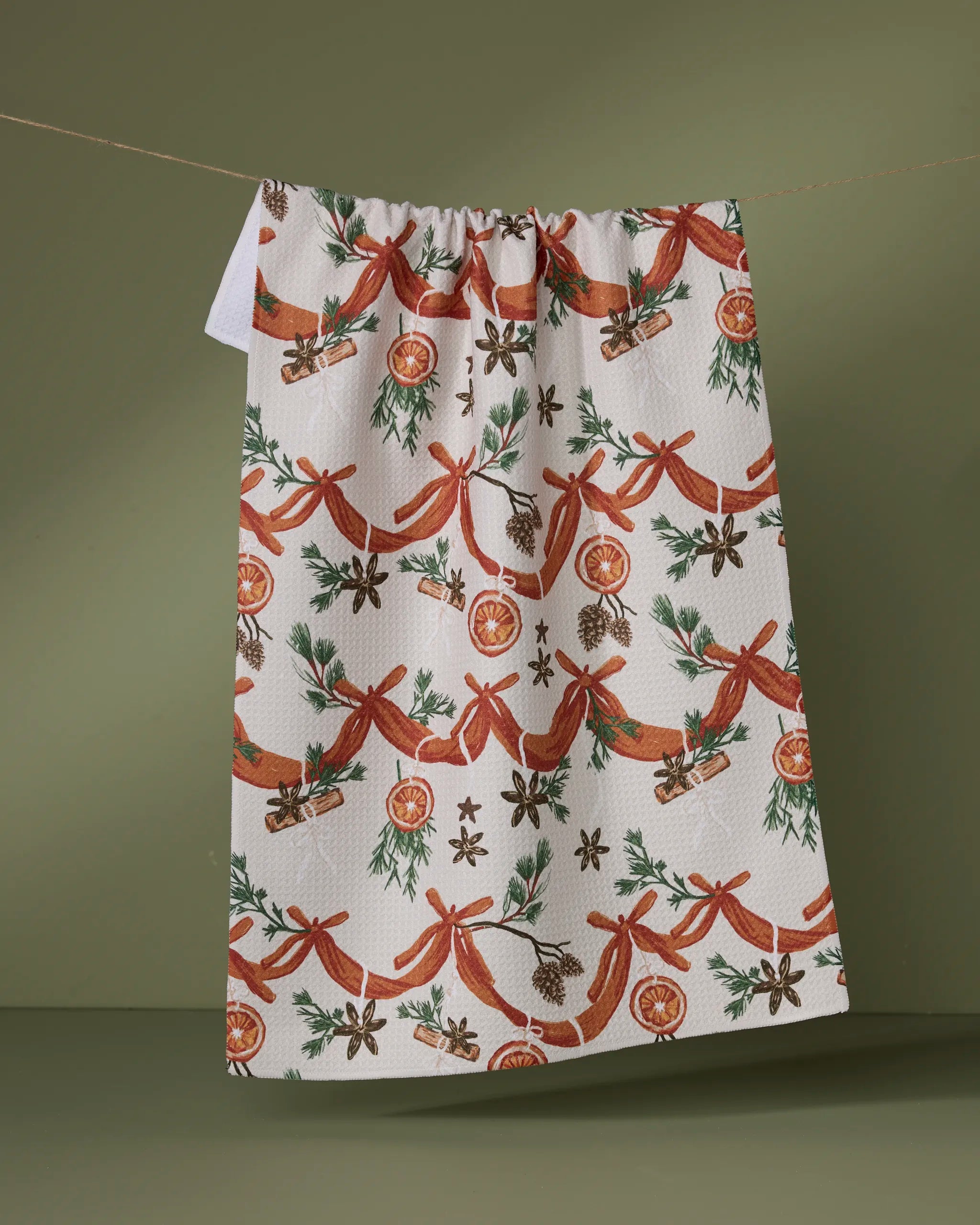 The Geometry Naranjas tea towel features a festive pattern of red bows, dried oranges, cinnamon sticks, pinecones, star anise, and green foliage, hanging on a line against an olive green background.