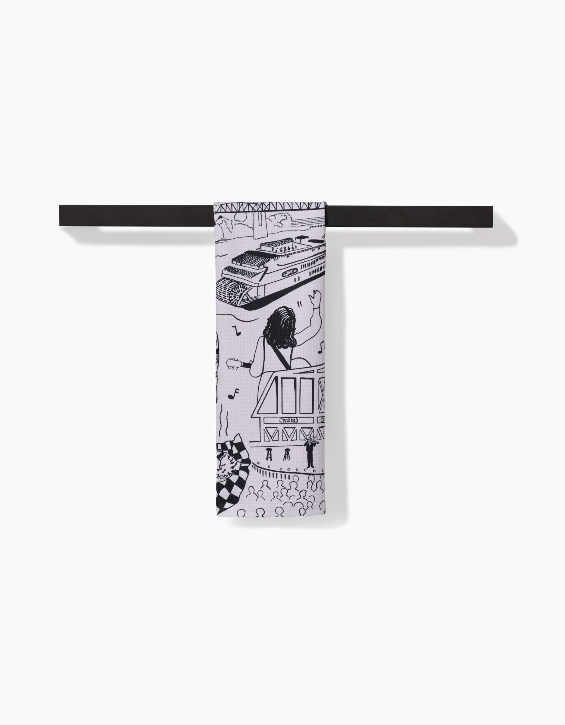 The Nashville tea towel by Elisabeth Wing features black line drawings of a tank, woman, house, and abstract patterns on white Geoweave™ fabric. It is draped over a minimalist dark bar set against a white background.
