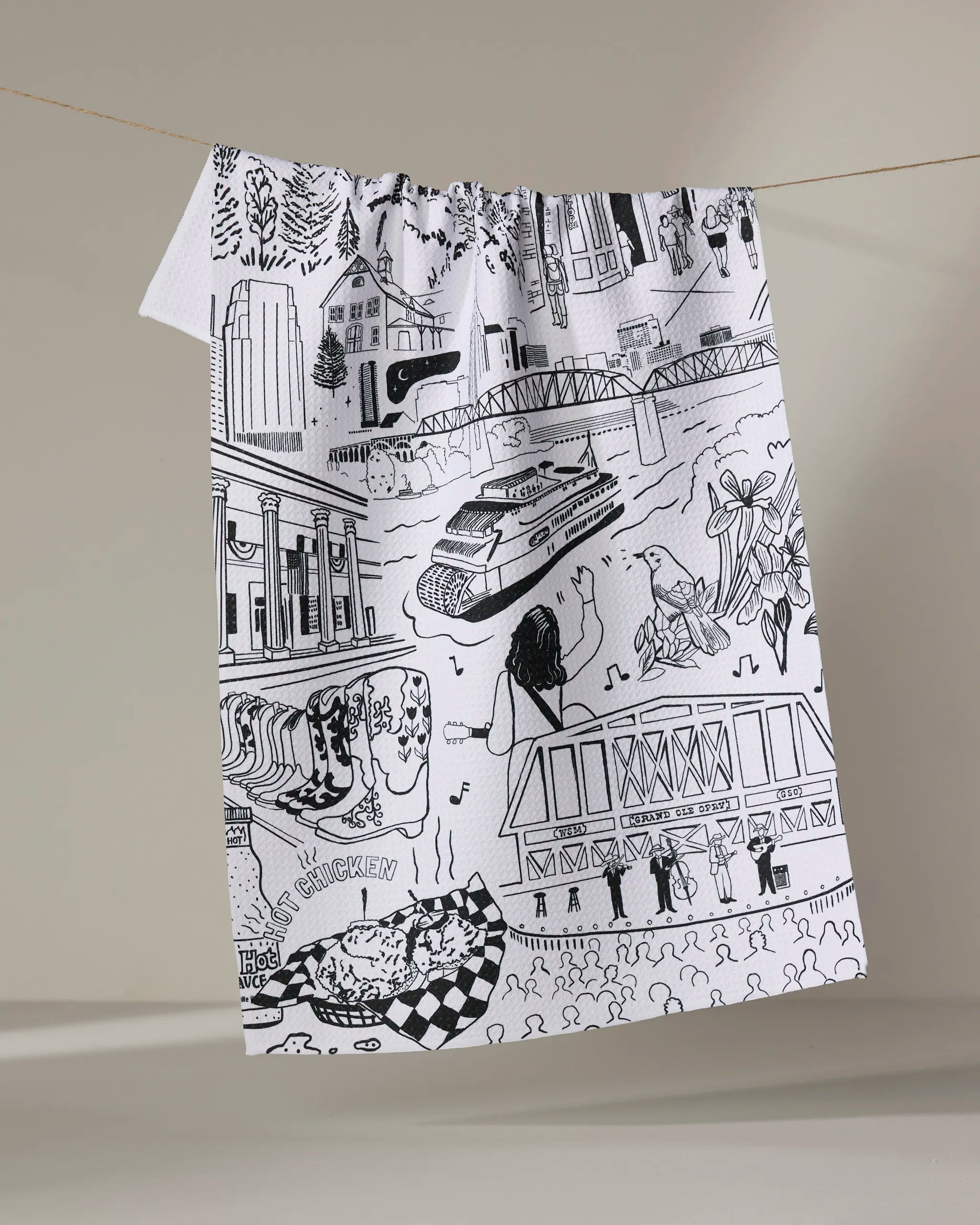 The Elisabeth Wing "Nashville" kitchen tea towel features black line drawings of the city's buildings, riverboat, bridge, people, birds, plants, and food. Made from recycled materials and ultra-absorbent, it hangs stylishly against a plain backdrop.