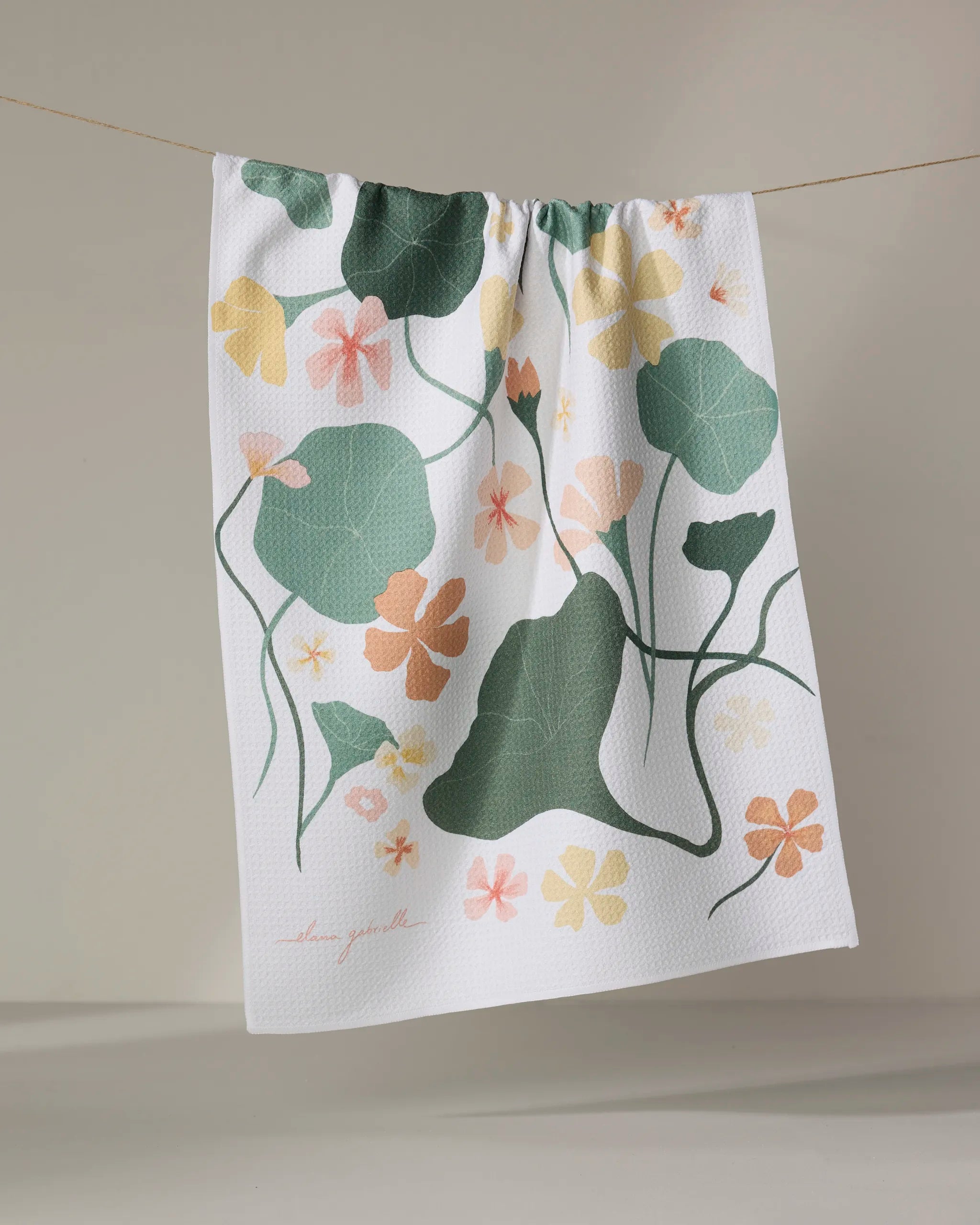 A recycled kitchen towel, Nasturtiums by Elana Gabrielle, hangs on a line with a botanical design of green leaves and orange, peach, and yellow flowers on a light background.