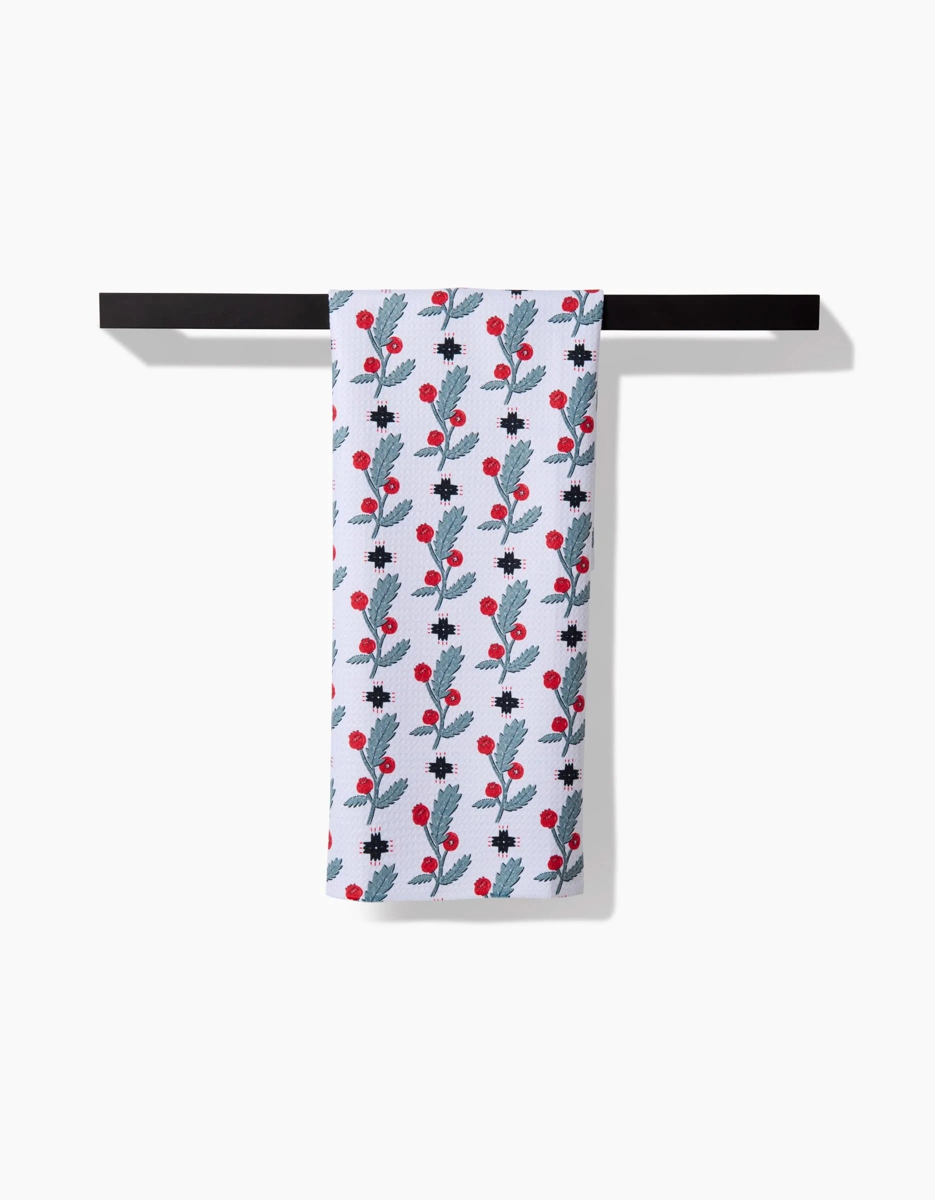 The Roxy Marj Native Holly tea towel, featuring a white background with blue leaves and red berries, hangs neatly on a black rack against a plain white wall.