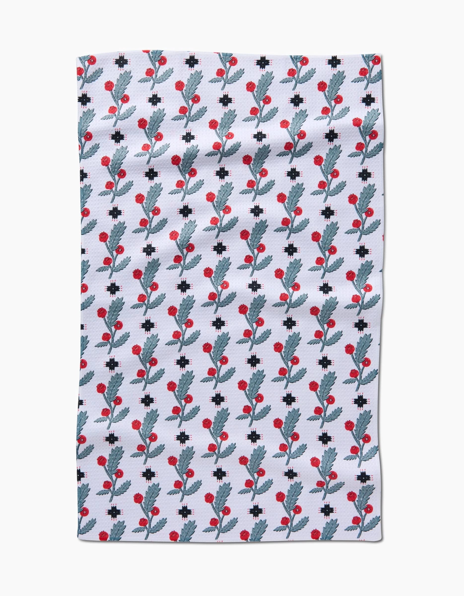 The Native Holly tea towel by Roxy Marj is rectangular and white, featuring vertical rows of blue leaves and red berries with scattered small black crosses.