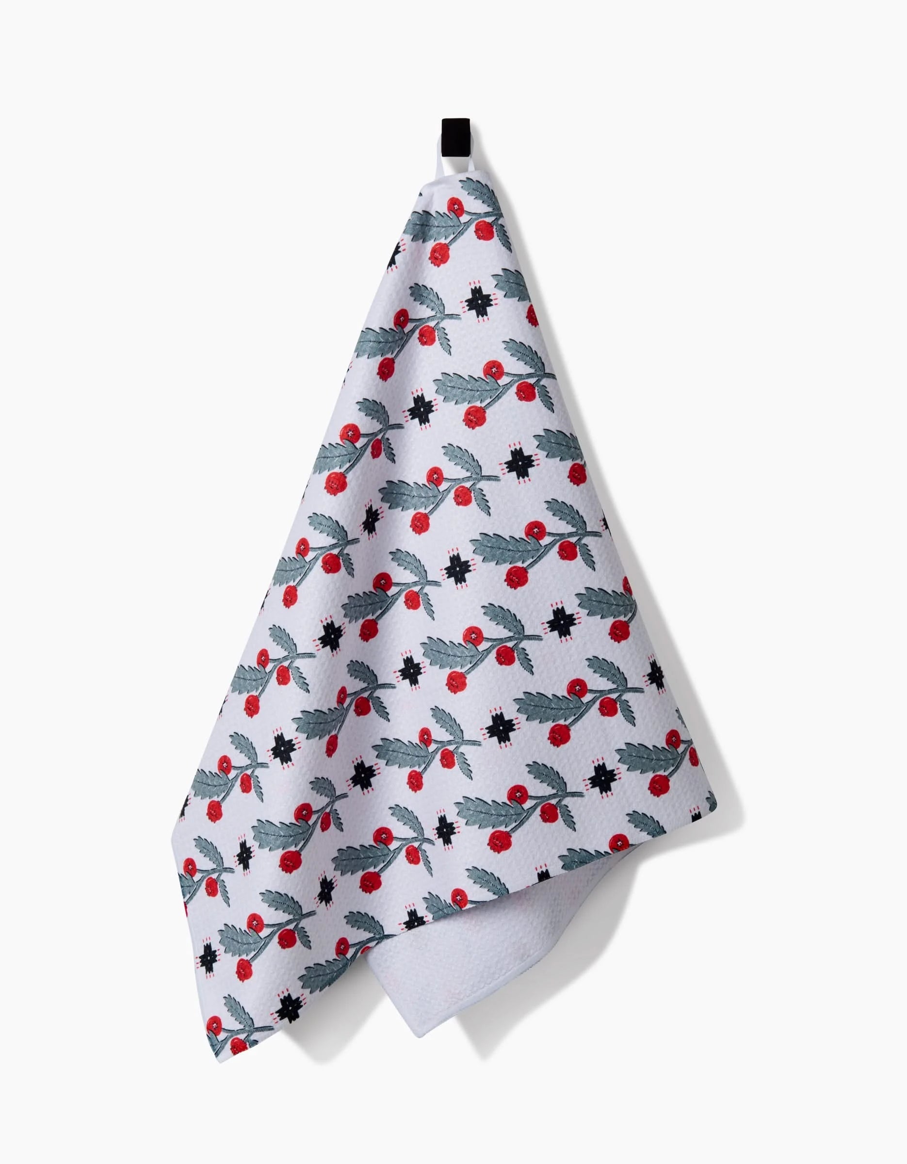 The Native Holly tea towel by Roxy Marj features green holly leaves, red berries, and black stars on white. Hanging by a black loop, it’s a festive and eco-friendly addition to any kitchen.