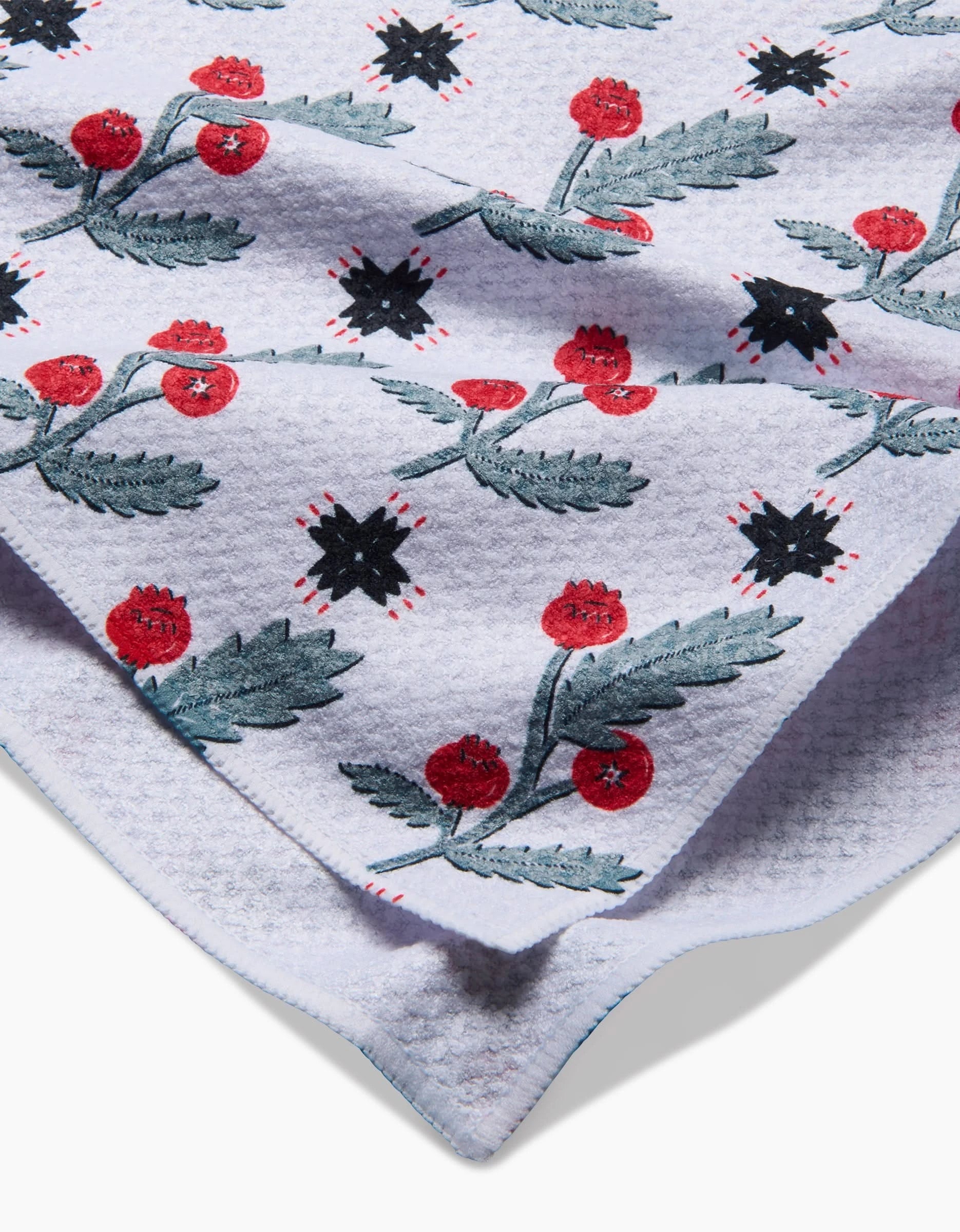 A close-up of the ultra-absorbent Roxy Marj Native Holly tea towel, light purple with red berries, green leaves, and black star shapes. The folded towel shows both front and back textured designs.