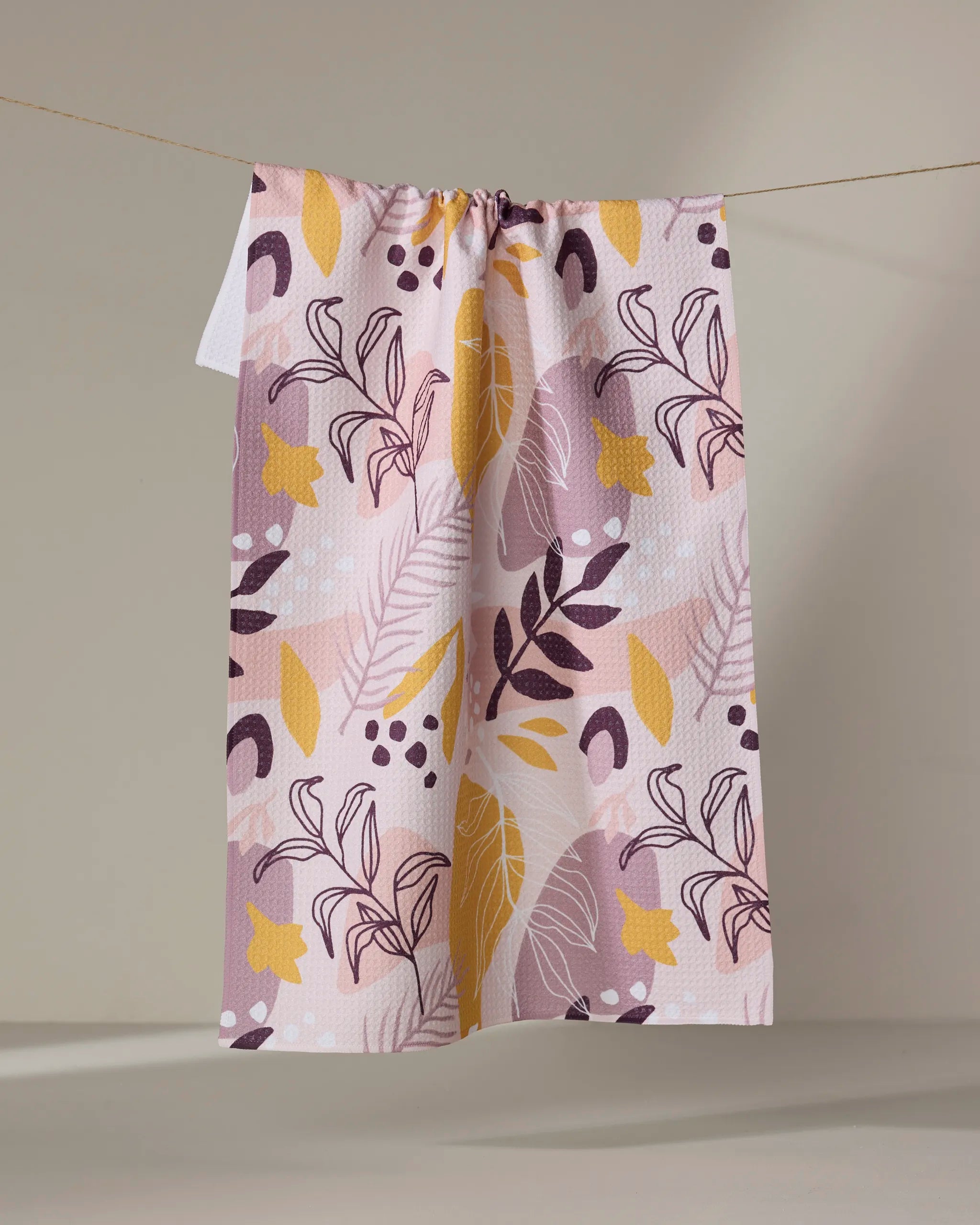 The Cottonflower Studio "Nature Sketch" tea towel, featuring a pink, yellow, and purple abstract leafy design, hangs on a clothesline against a neutral beige background.