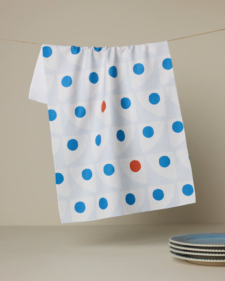The Geometry "Nautical Eye" recycled kitchen towel hangs on a string, featuring blue dots with one red dot. Below, three stacked blue and white plates rest on a light surface.