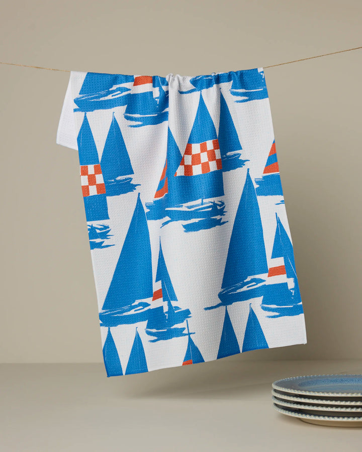 The Geometry Nautical Mile tea towel, featuring blue sailboats with red checkered sails, hangs on a string above a stack of blue-rimmed plates against a beige background.