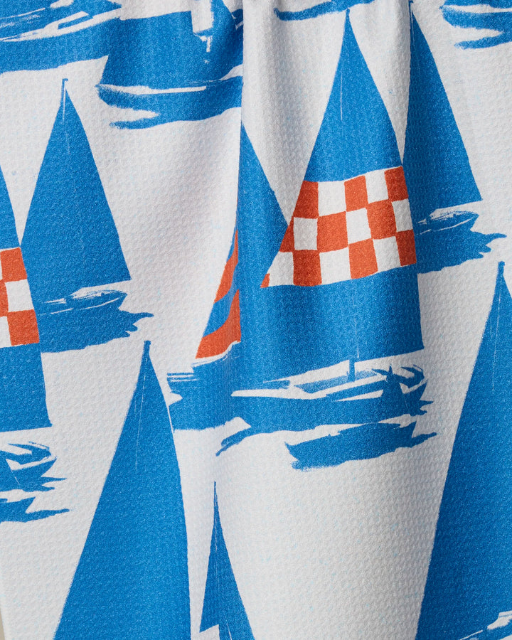 The Geometry "Nautical Mile" tea towel showcases blue sailboats with red and white checkered sails on a white background accented by blue brushstroke waves.