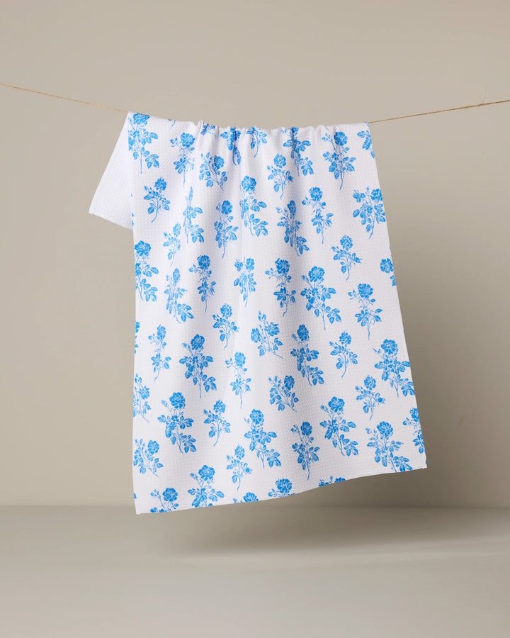 The Geometry Naxos tea towel, featuring a blue floral pattern, hangs from a string against a plain beige background.