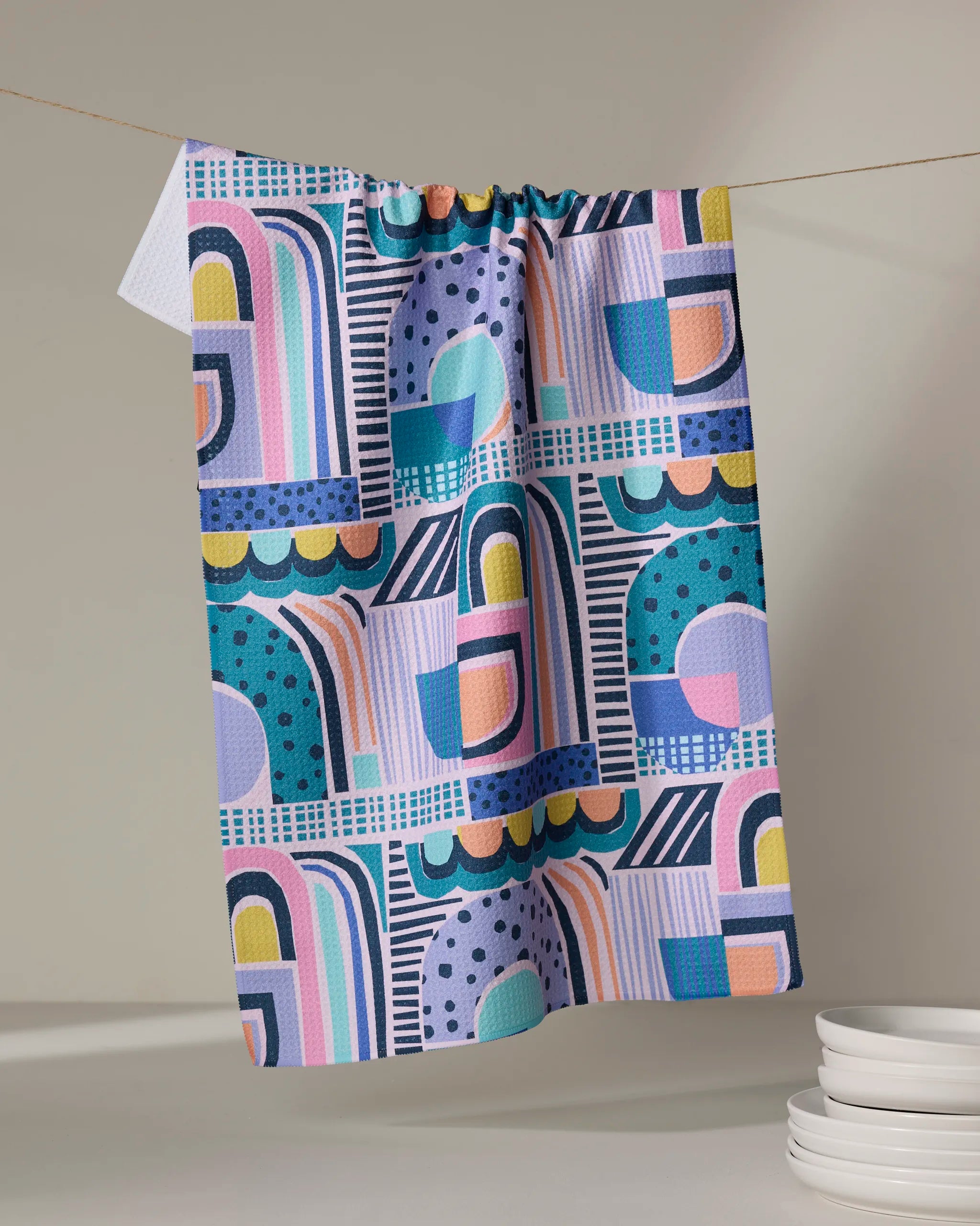 The New Dawn tea towel by Masha Volnova, made from recycled materials, displays a bold geometric pattern in blue, pink, purple, and yellow. It hangs on a line next to stacked white plates on a neutral surface.