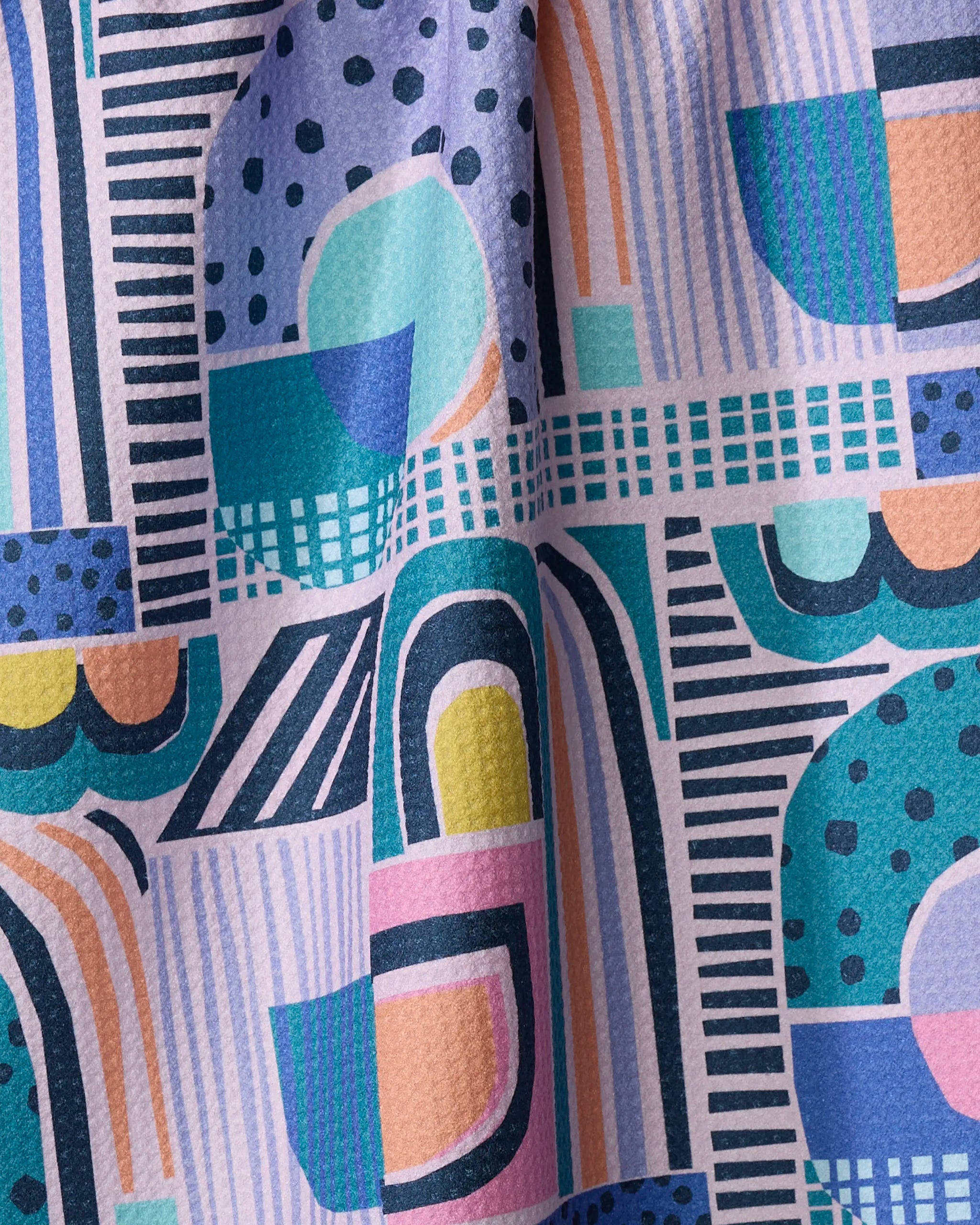 Close-up of the New Dawn kitchen tea towel by Masha Volnova, made from Geoweave fabric with geometric patterns in bold blue, teal, orange, purple, and pink. The colorful fabric is slightly wrinkled or folded.